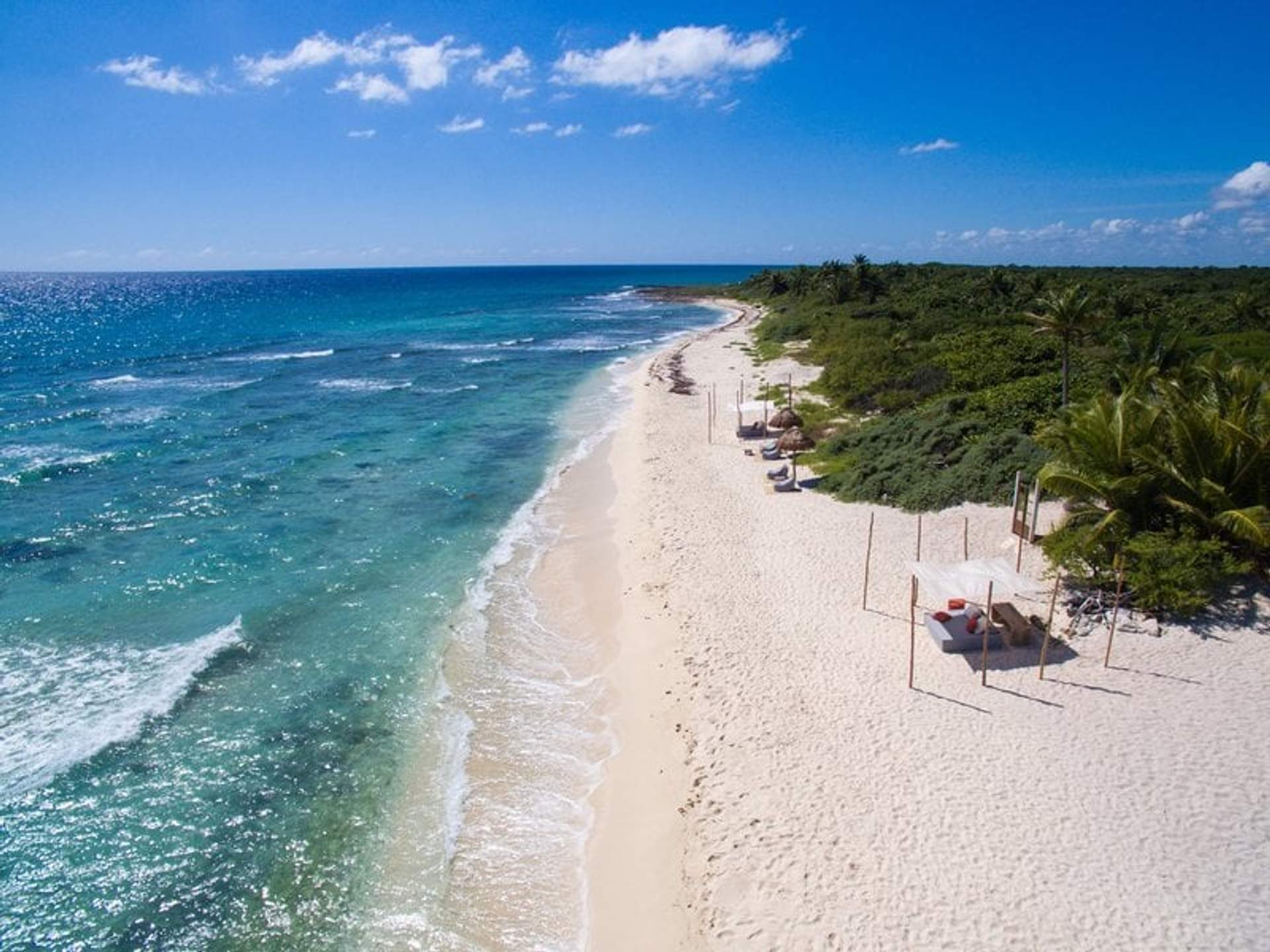 Vista aérea de una playa de arena blanca y agua turquesa con varias cabañas y áreas de descanso a lo largo de la orilla, rodeada de vegetación tropical bajo un cielo despejado.