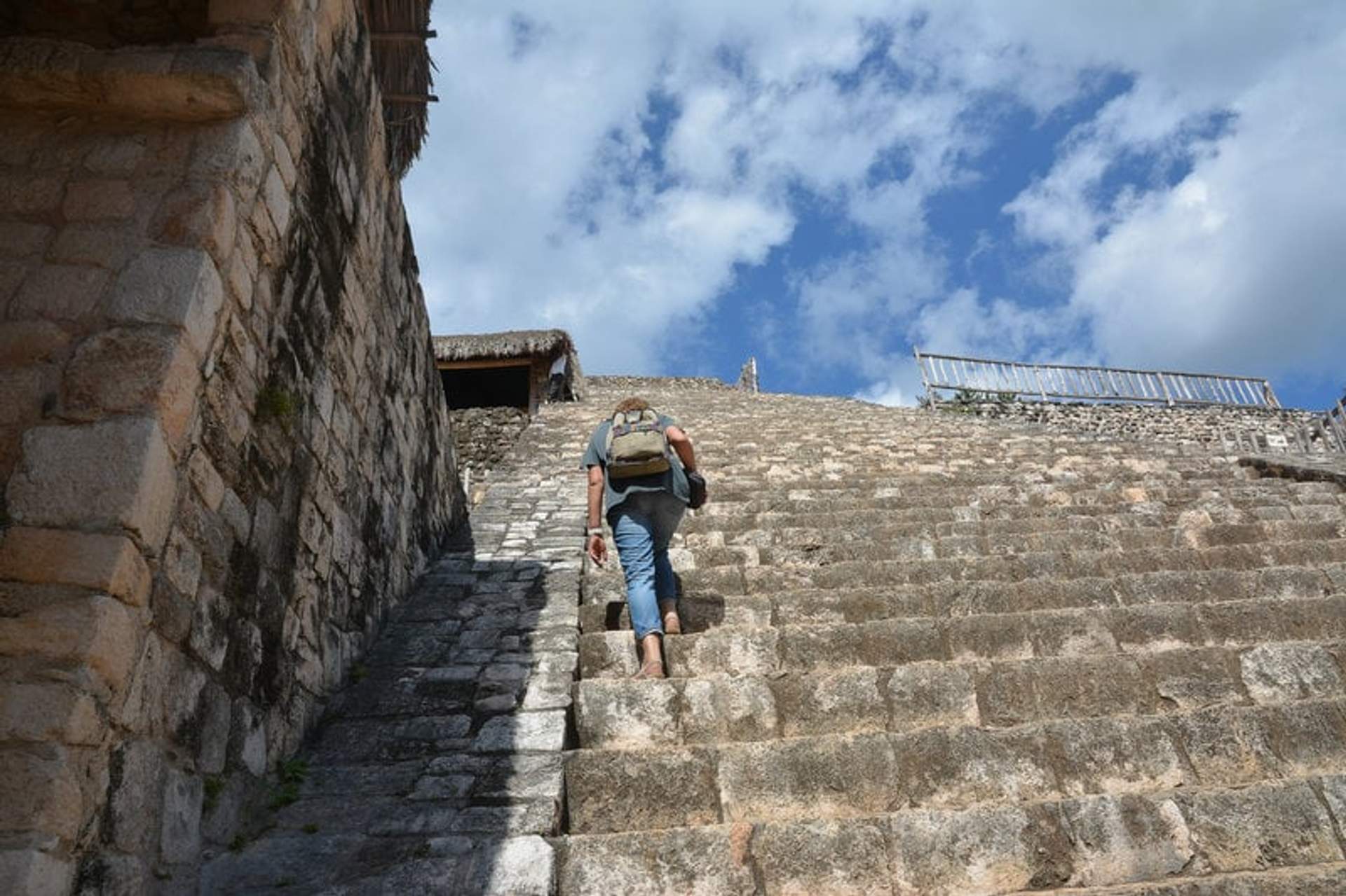 A person climbing the steep stone steps of a pyramid at Ek Balam, with a blue sky and scattered clouds overhead.