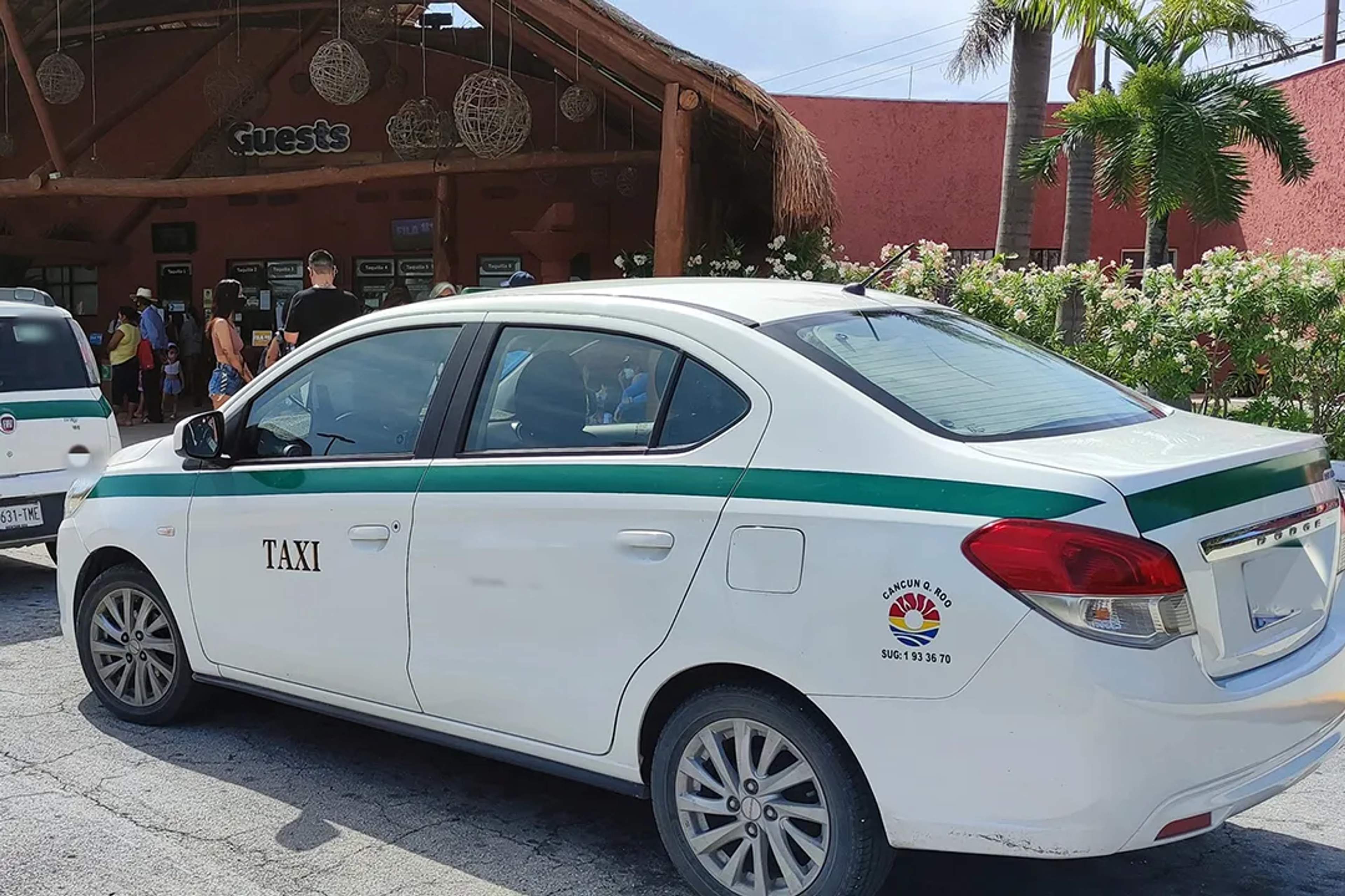 White Cancun taxi with green stripe parked near a tropical entrance filled with tourists.