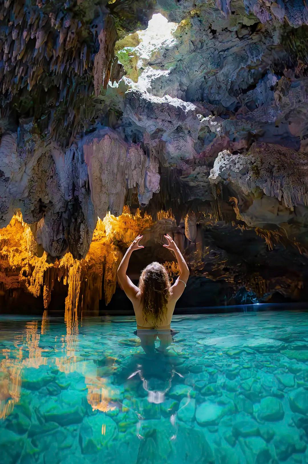 A woman stands in a crystal-clear cenote, reaching towards the light streaming through the cave's ceiling.