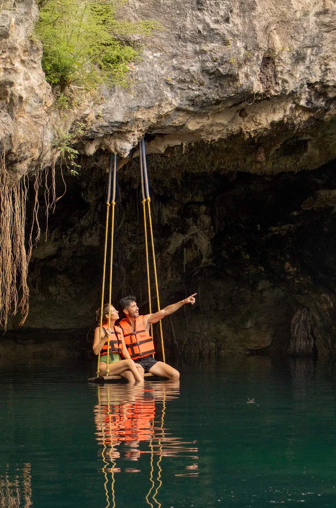 A couple in life jackets sits on a swing above a cenote, admiring the beauty of the cave and its waters.