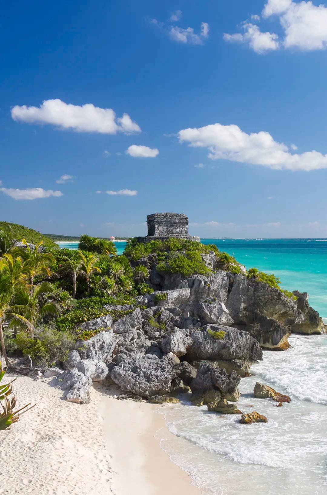 A rocky cliff with ancient ruins overlooks a pristine beach and turquoise waters under a bright, clear sky in Tulum.
