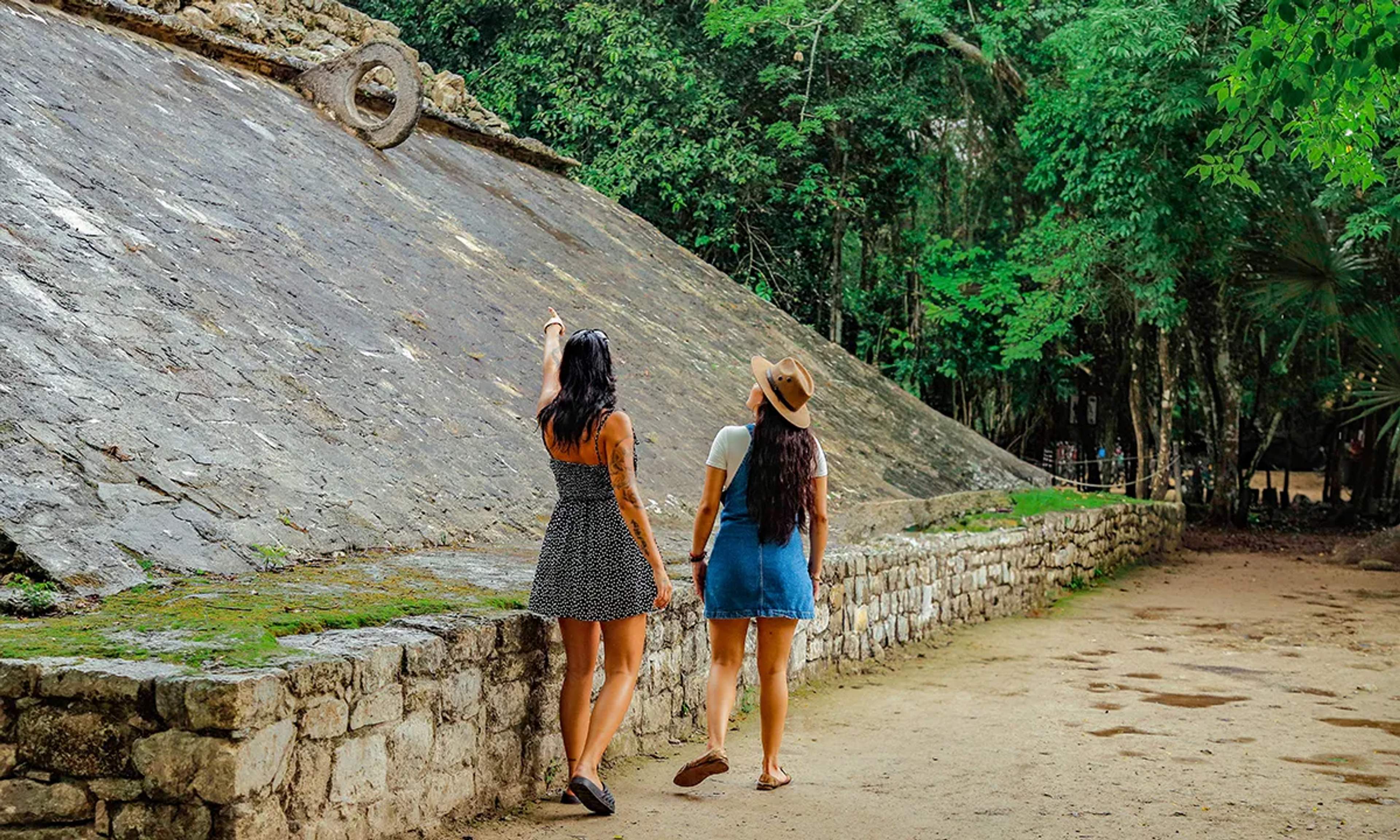 Two women walking by an ancient stone ball court, one pointing at the stone hoop, surrounded by jungle vegetation.