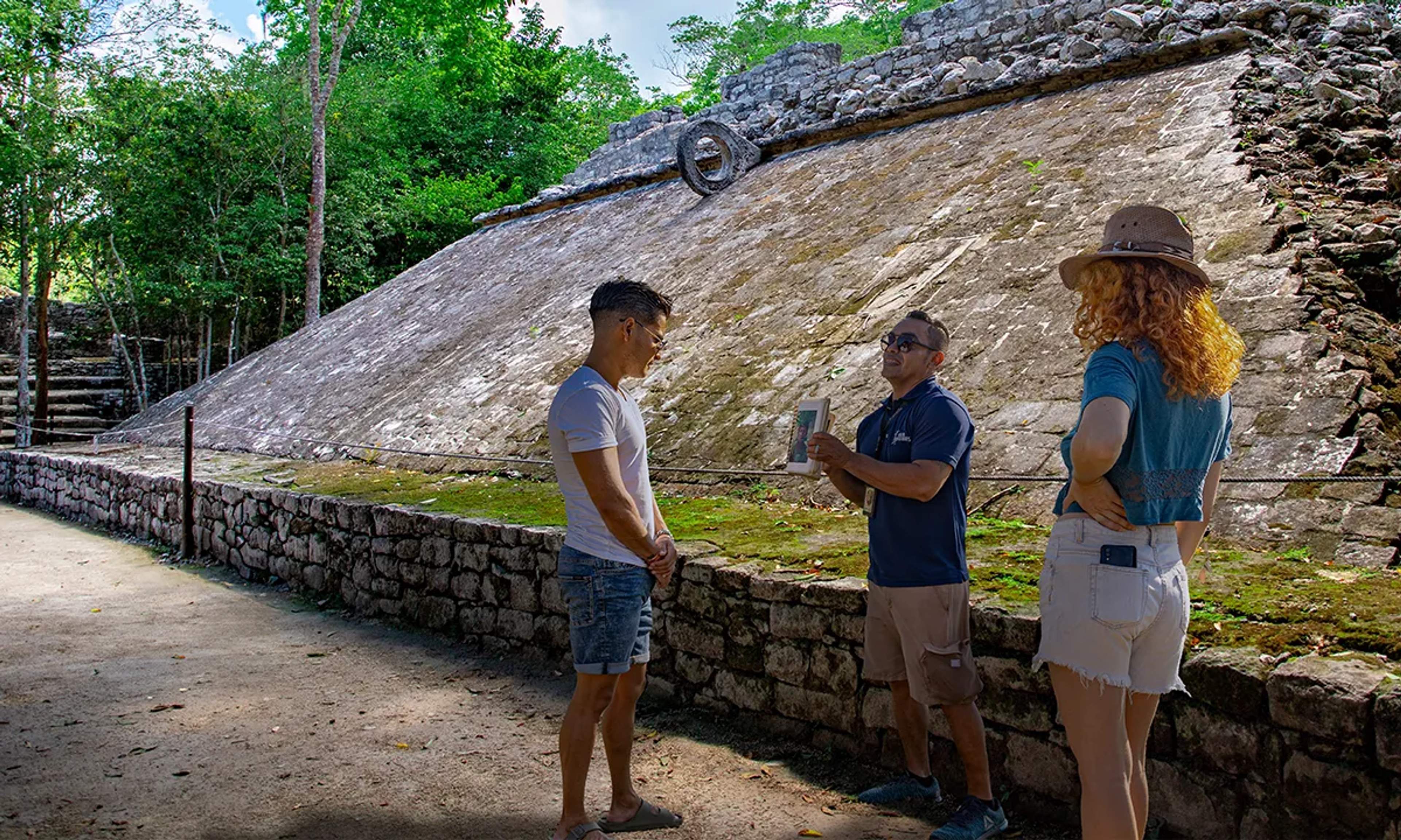 A guide explains the ancient ball court to two visitors, standing by the sloped stone structure surrounded by jungle.