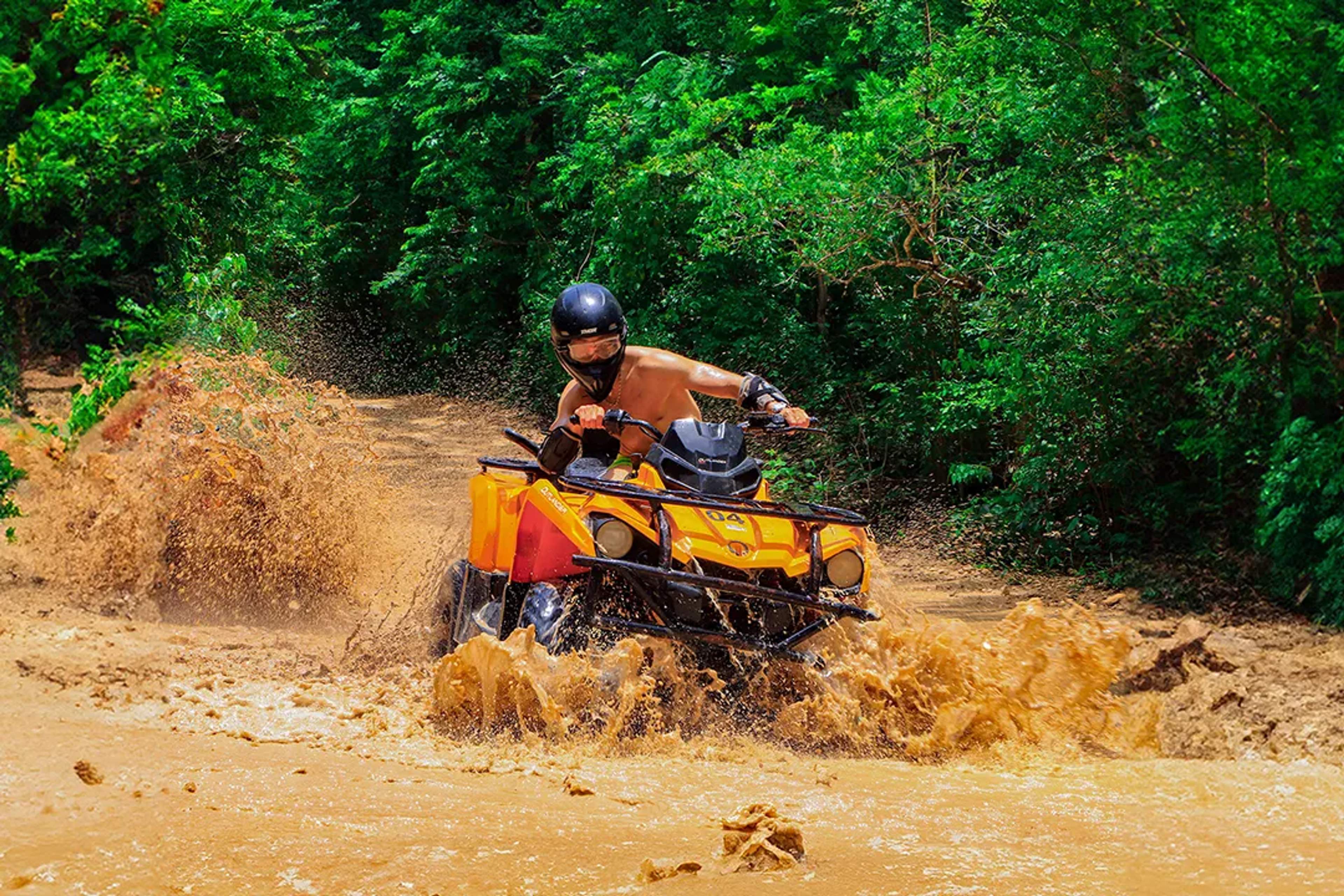 Rider on an ATV splashes through a muddy jungle trail, surrounded by lush green trees.