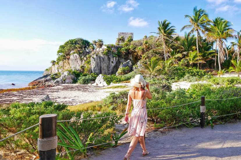 Mujer con vestido veraniego admirando las ruinas de Tulum junto al mar.