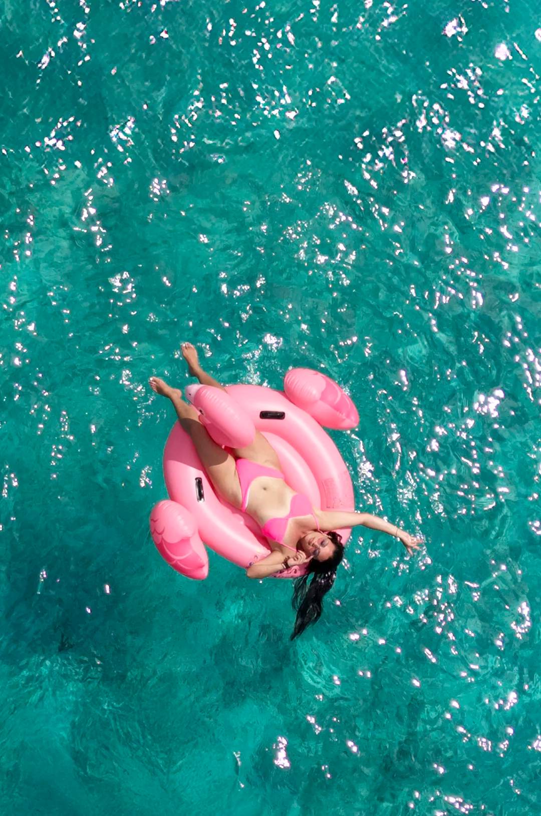 A woman in a pink bikini relaxes on a pink flamingo float in crystal-clear turquoise waters.