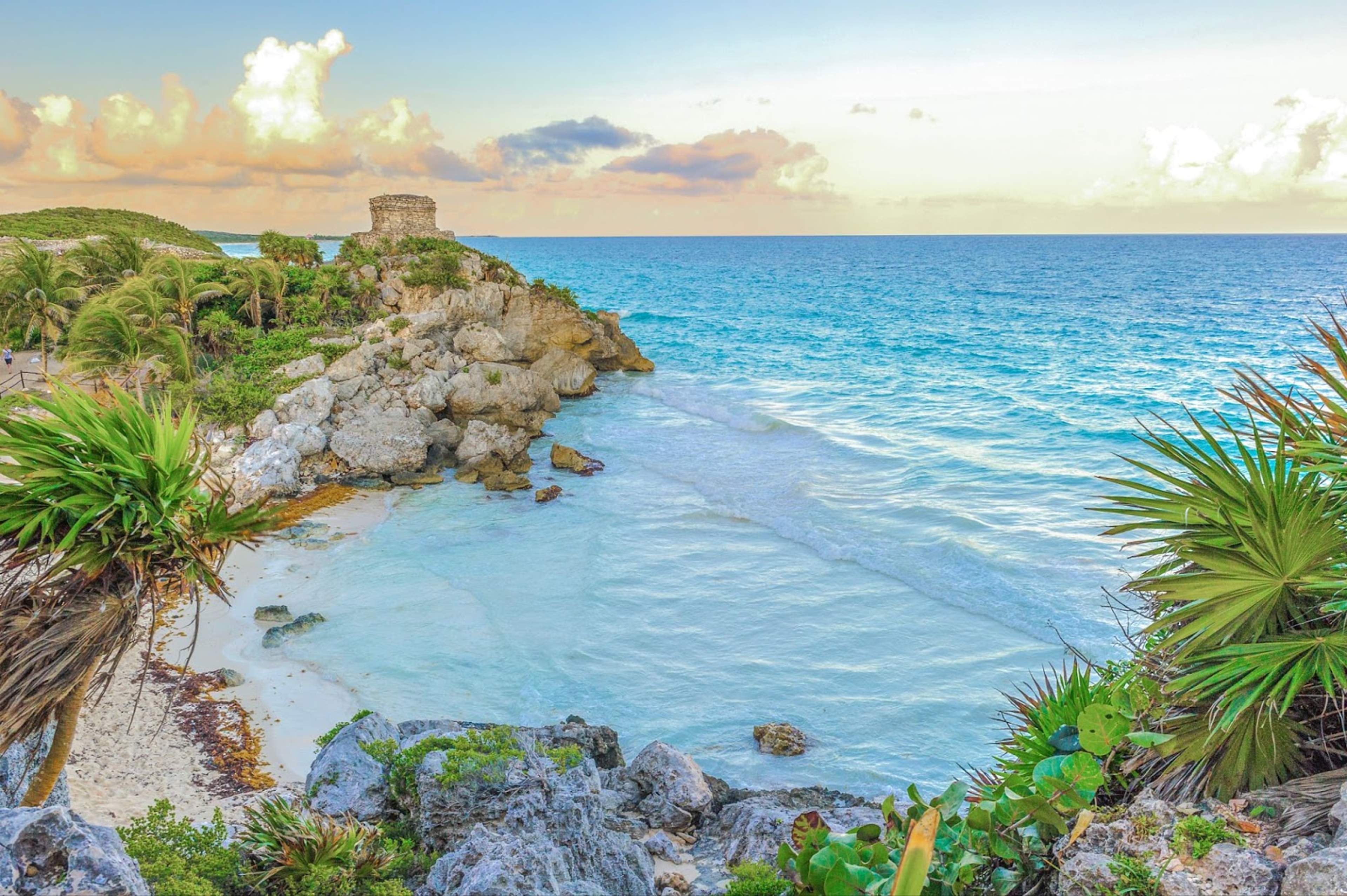 A scenic view of Tulum's coastline featuring ancient ruins perched on a cliff, overlooking turquoise waters and a sandy beach.