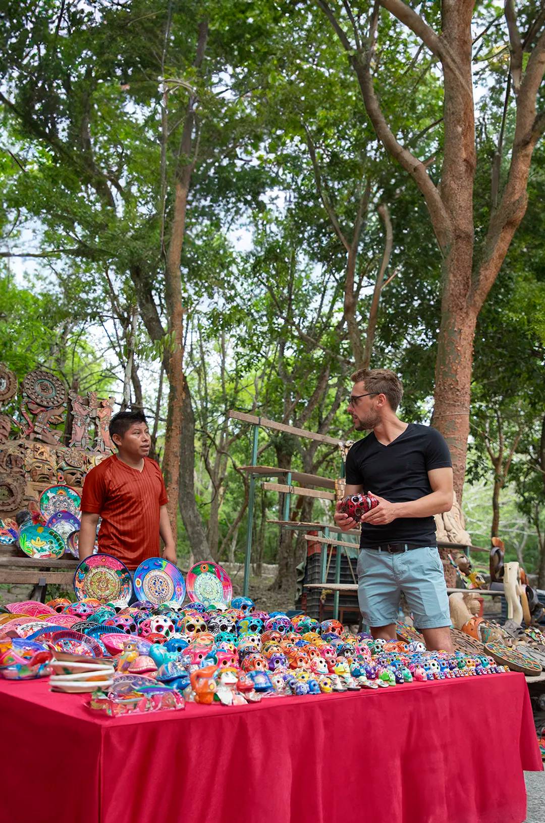 A tourist shops for colorful handcrafted souvenirs at an outdoor market in Chichen Itza, surrounded by lush greenery.