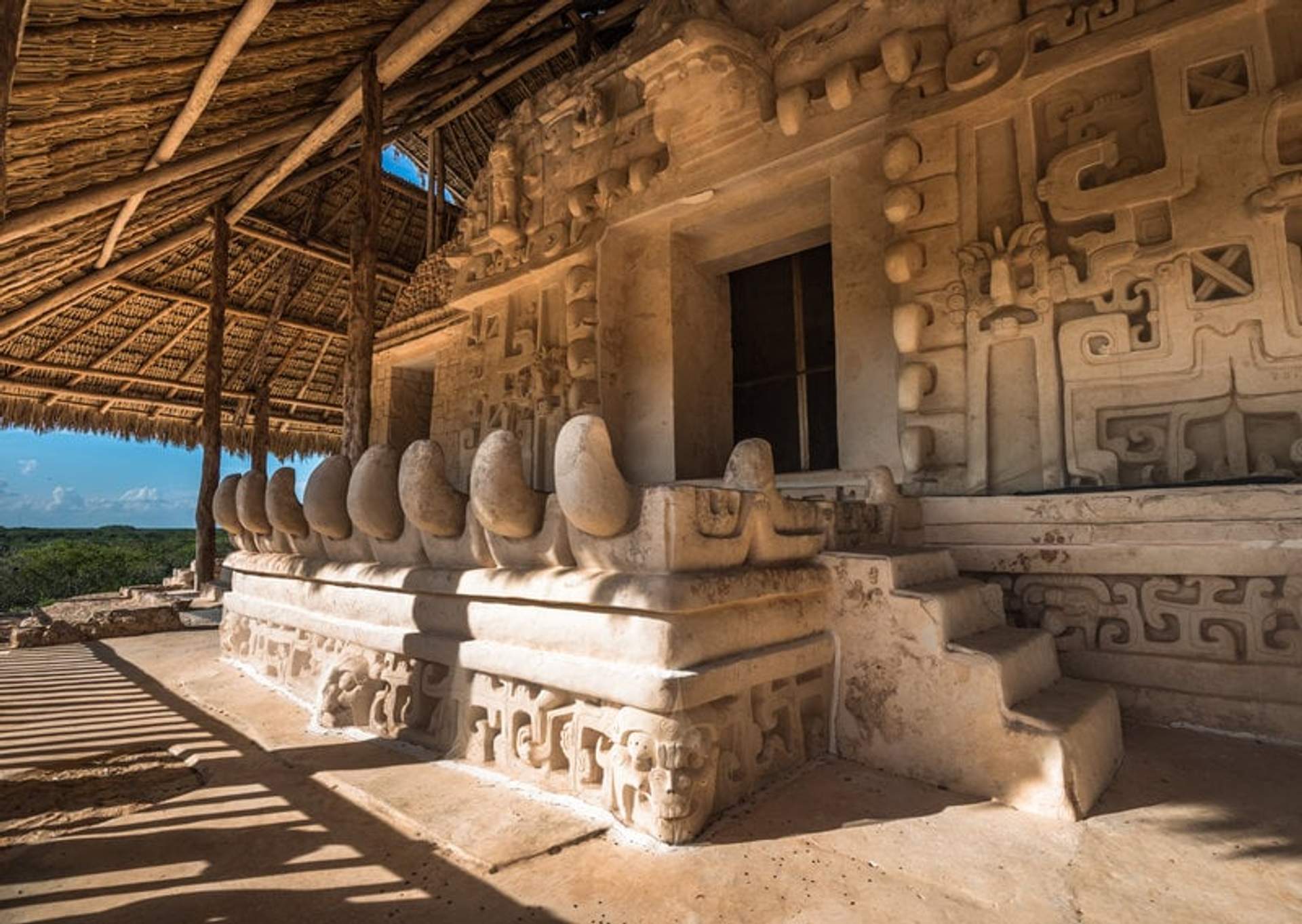 Close-up of intricate Mayan carvings and sculptures on a building at Ek Balam, partially shaded by a thatched roof.