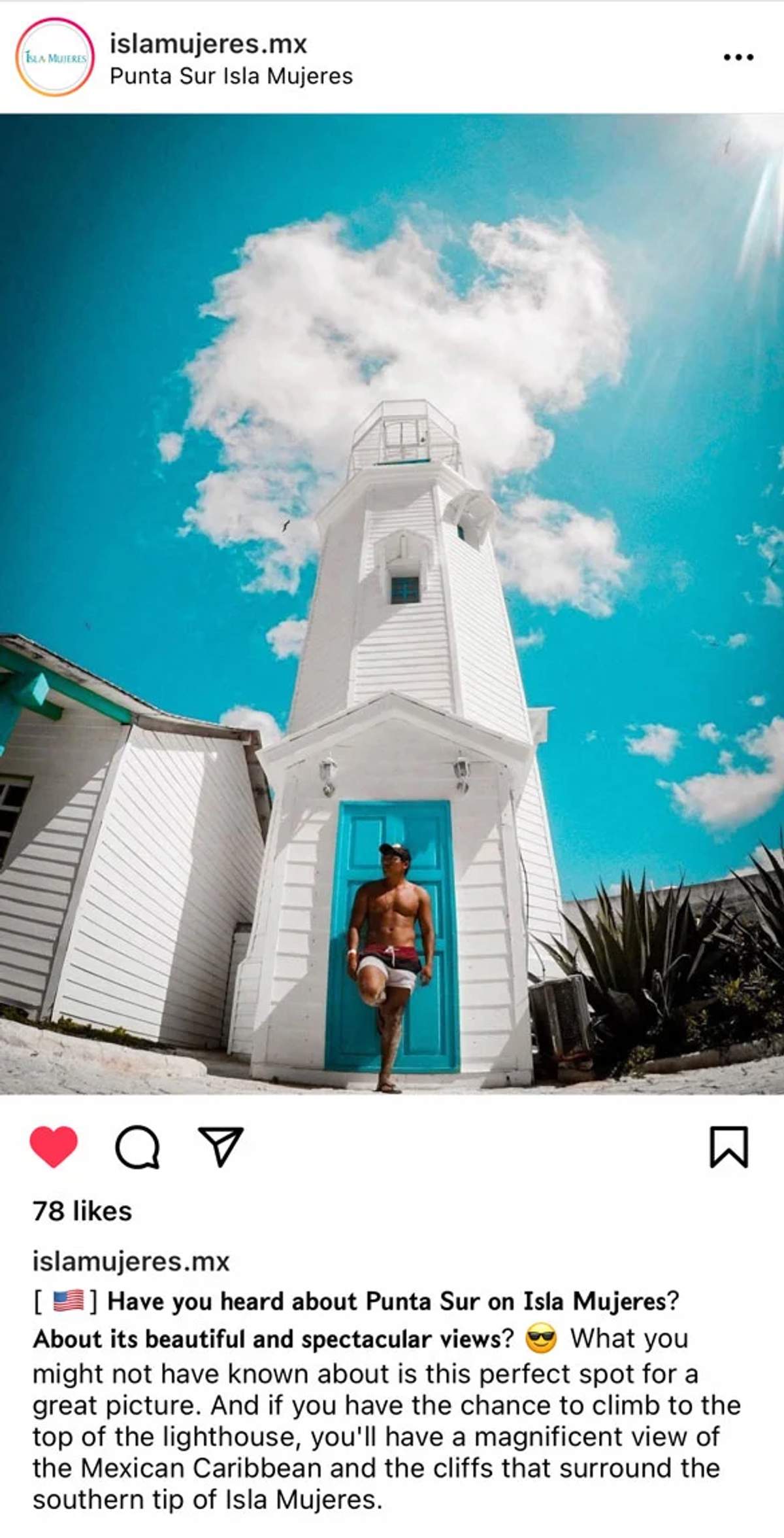 Un hombre sin camisa con shorts y gorra está de pie frente a un faro blanco con una puerta azul en Punta Sur, Isla Mujeres, bajo un cielo azul brillante con nubes esponjosas.
