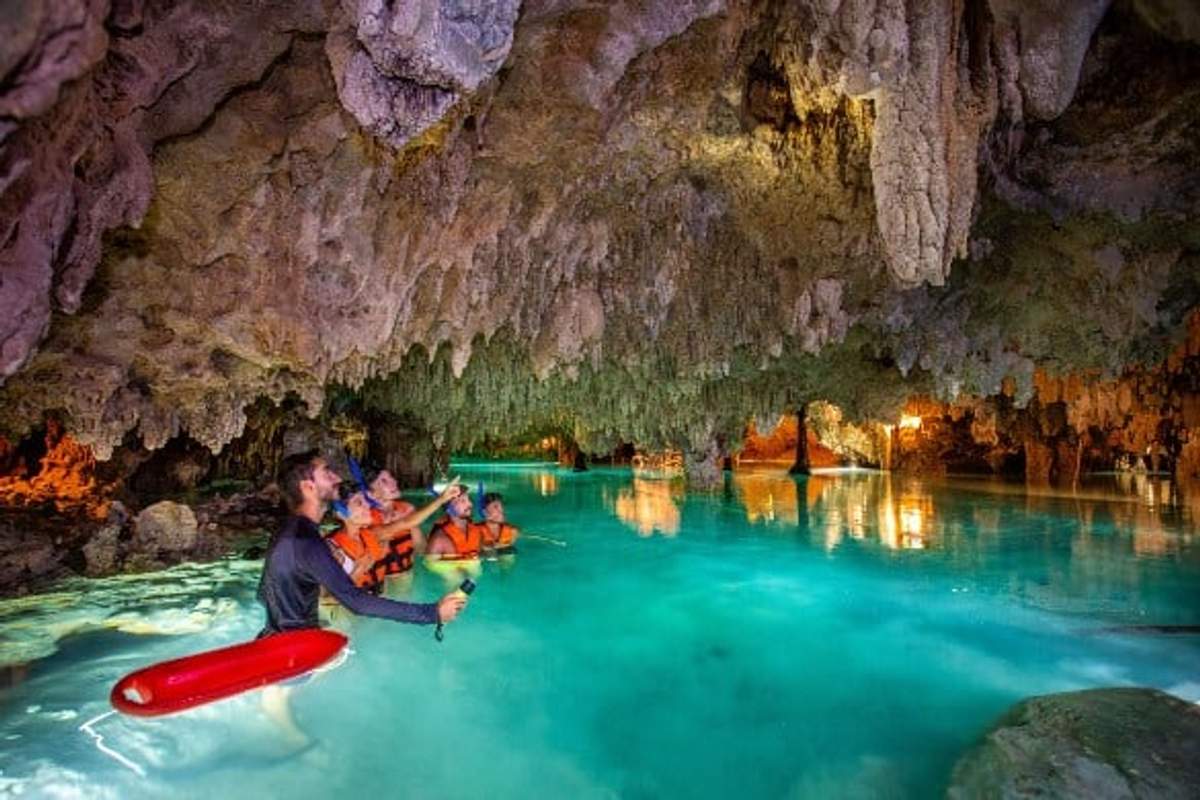Group of people exploring a cenote in Mexico, wearing life jackets and helmets, surrounded by stalactites and clear blue water.