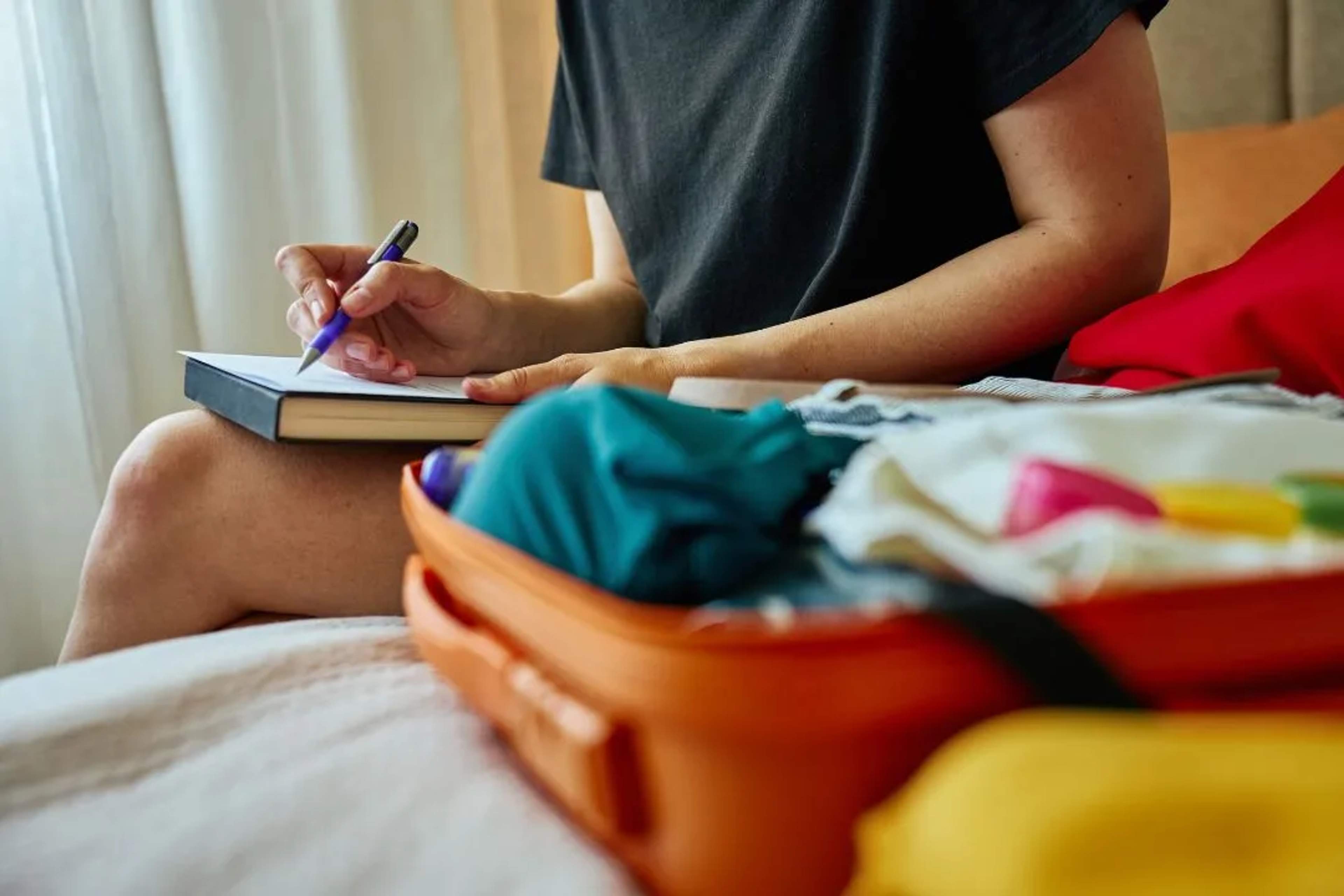 Persona tachando elementos de una lista de equipaje en la cama con una maleta abierta.