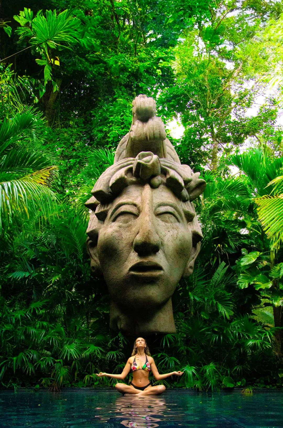 Woman meditating in a photogenic spot in Tulum, with a Mayan statue and lush greenery.