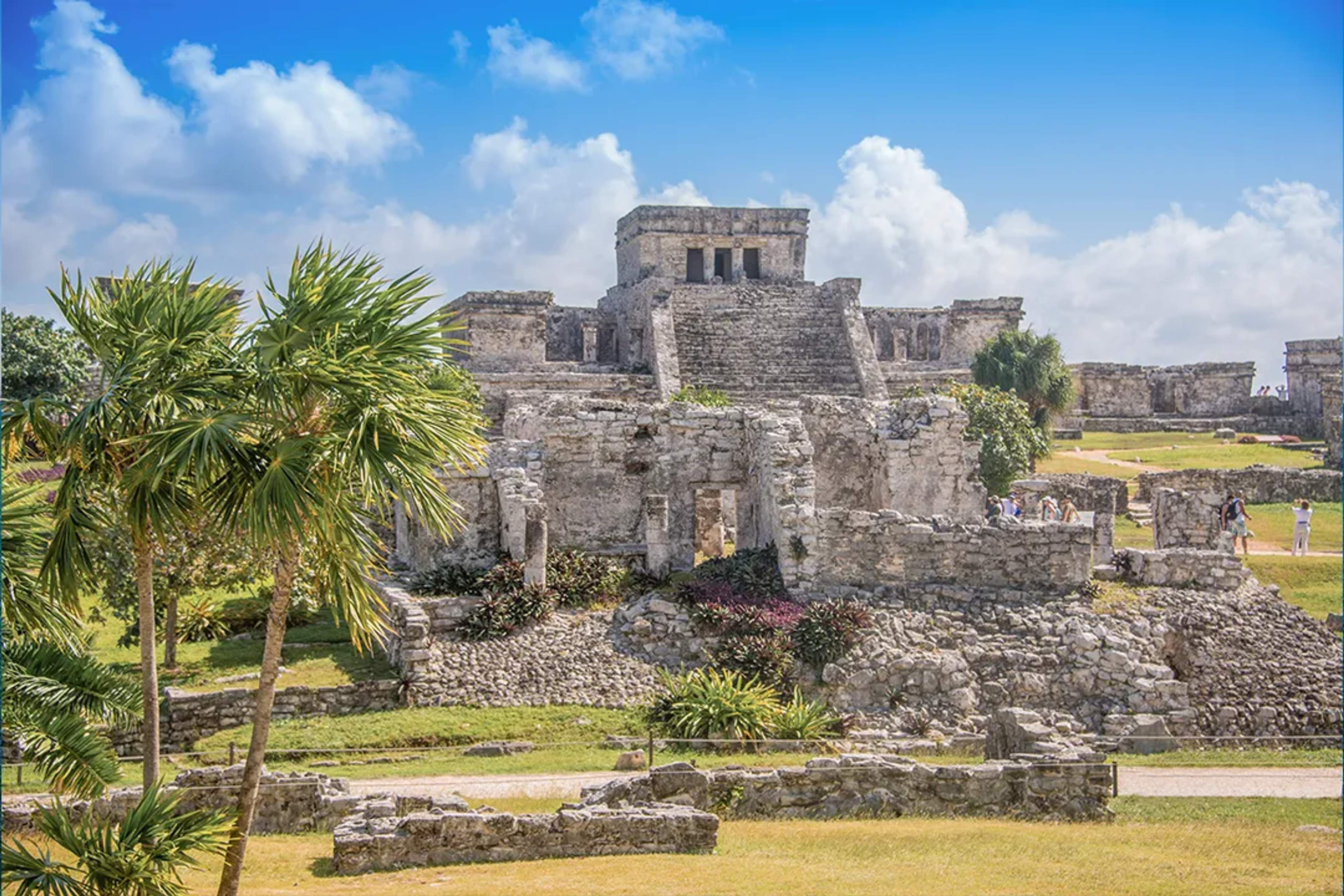 Antiguas ruinas mayas de Tulum con palmeras y turistas bajo un cielo azul brillante.