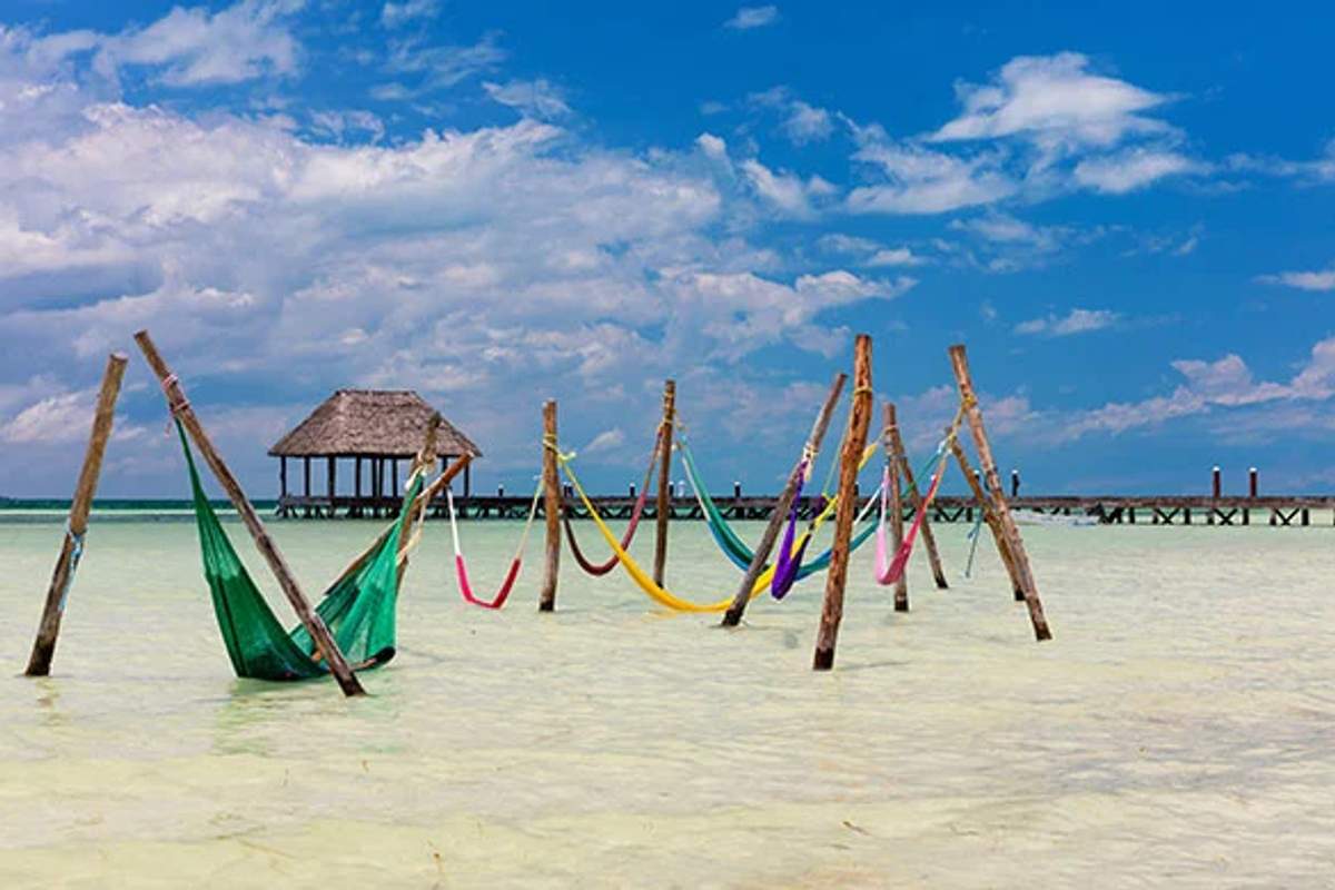 Hamacas coloridas cuelgan sobre aguas poco profundas en Holbox, con un muelle y una choza de fondo.