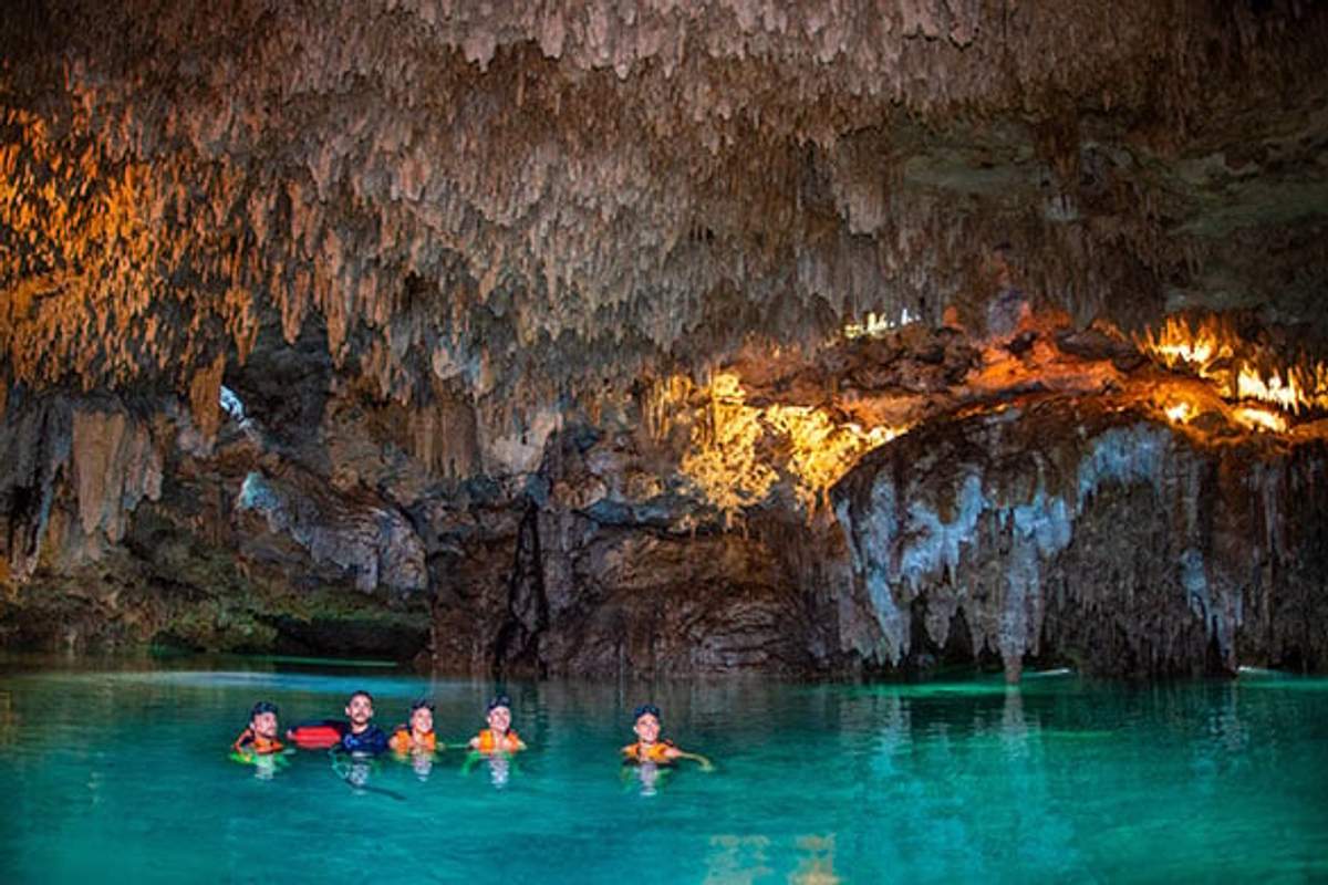 Un grupo de personas nadando en las aguas turquesas de un cenote en Cancún, rodeados de estalactitas iluminadas y paredes de cueva.