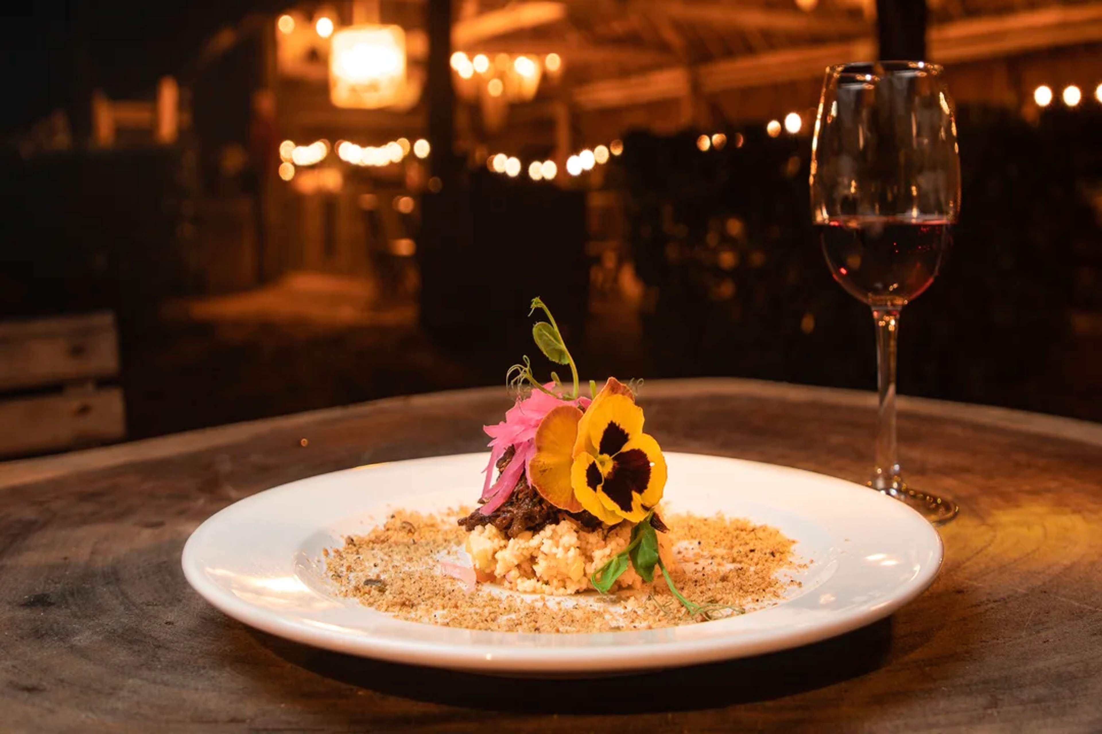 Plato elegante con flores comestibles y vino, en mesa rústica bajo luces cálidas nocturnas.