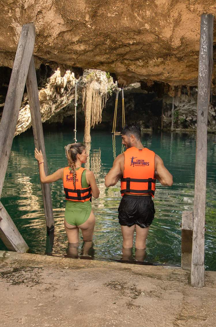 A couple wearing life jackets stands at the entrance of a cenote, ready to explore its crystal waters.d by stone walls