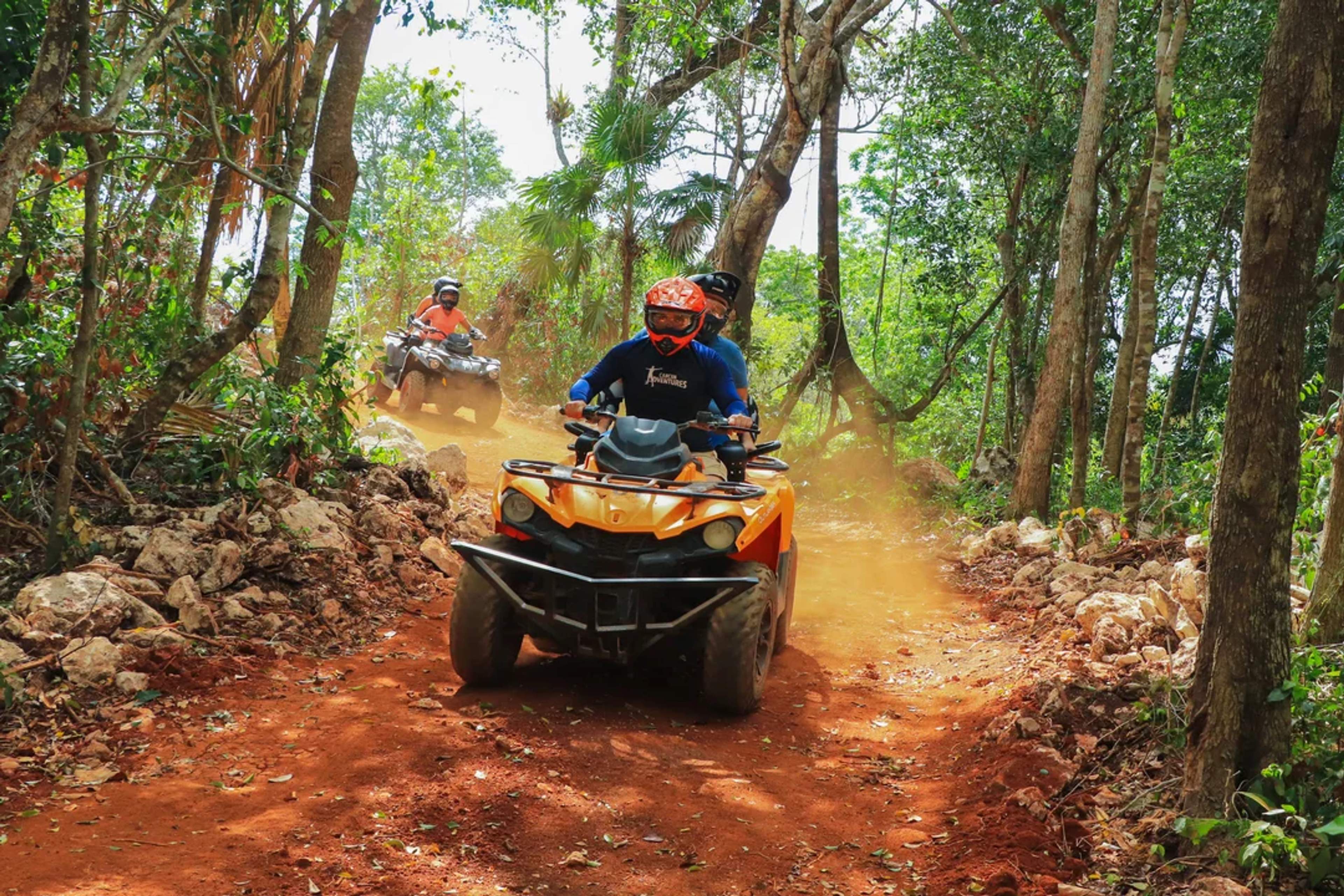 Aventureros conducen cuatrimotos por un sendero de tierra roja rodeado de selva, levantando polvo mientras exploran.