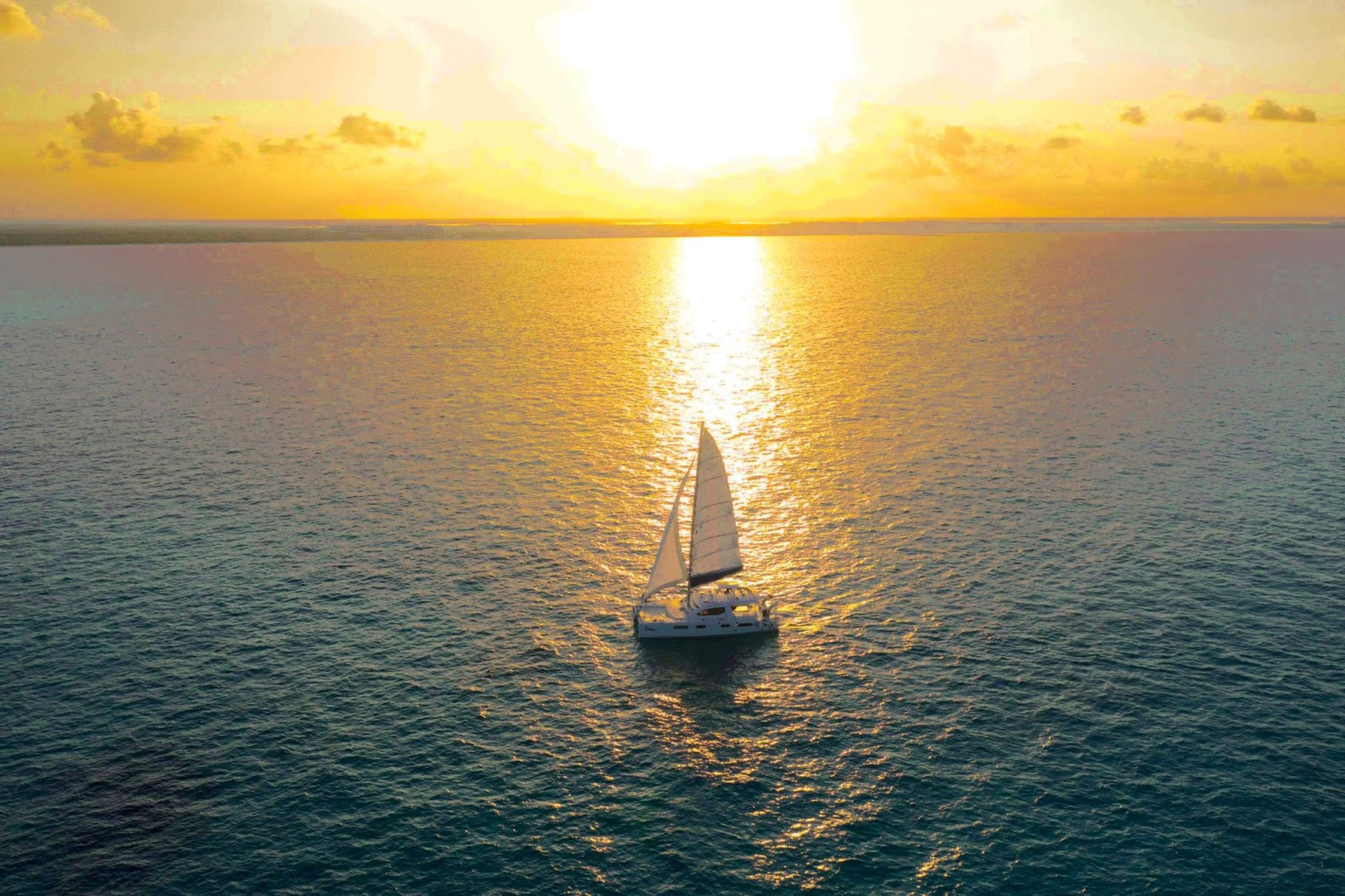 A sailboat glides across the ocean under a golden sunset, casting a warm reflection on the water.