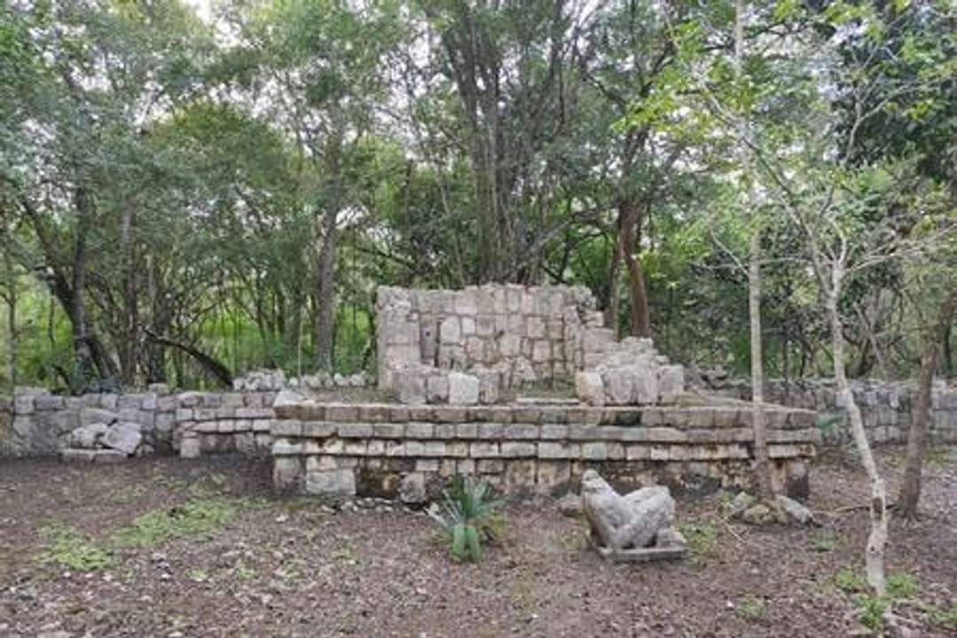 A photograph of the Casa Chac Mool at Chichen Itza, showcasing an ancient stone structure with a platform and partially ruined walls. The site is surrounded by dense trees and vegetation. Photo credit: Giovanni Agostino Frassetto.