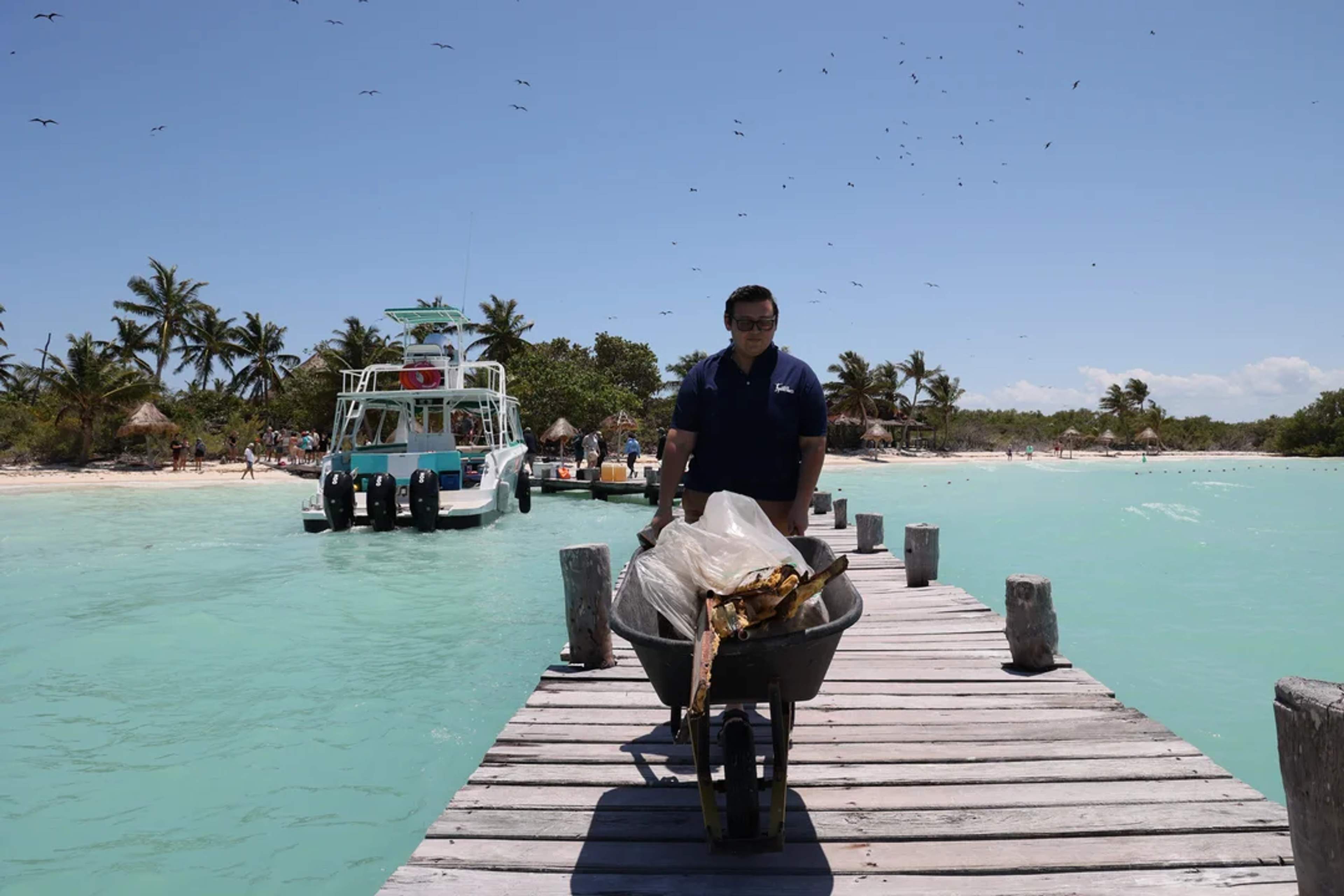 Man pushes a wheelbarrow full of trash on a dock after a beach cleanup near turquoise waters.
