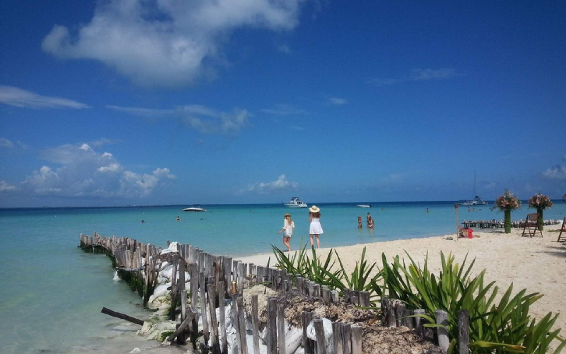 People walking on the sandy beach of Playa Norte, Isla Mujeres, with clear blue waters and boats in the distance.