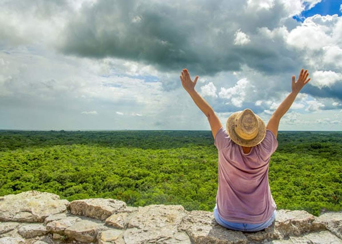 Una persona con un sombrero de paja y una camiseta rosa se sienta en unas ruinas antiguas con los brazos levantados, contemplando un vasto bosque verde bajo un cielo nublado y dramático.