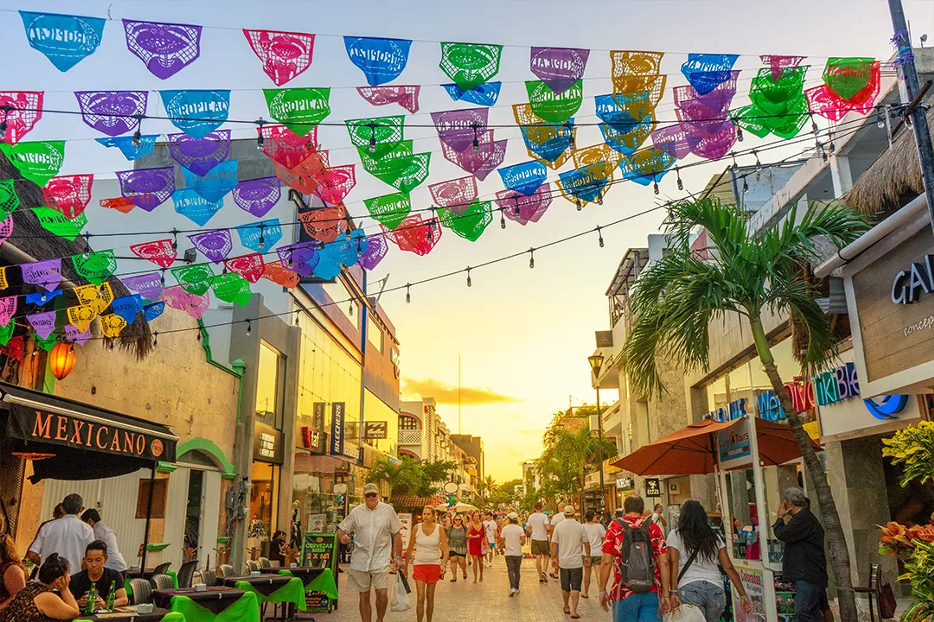 Banderas de colores sobre la 5ta Avenida en Playa del Carmen, llena de gente al atardecer.