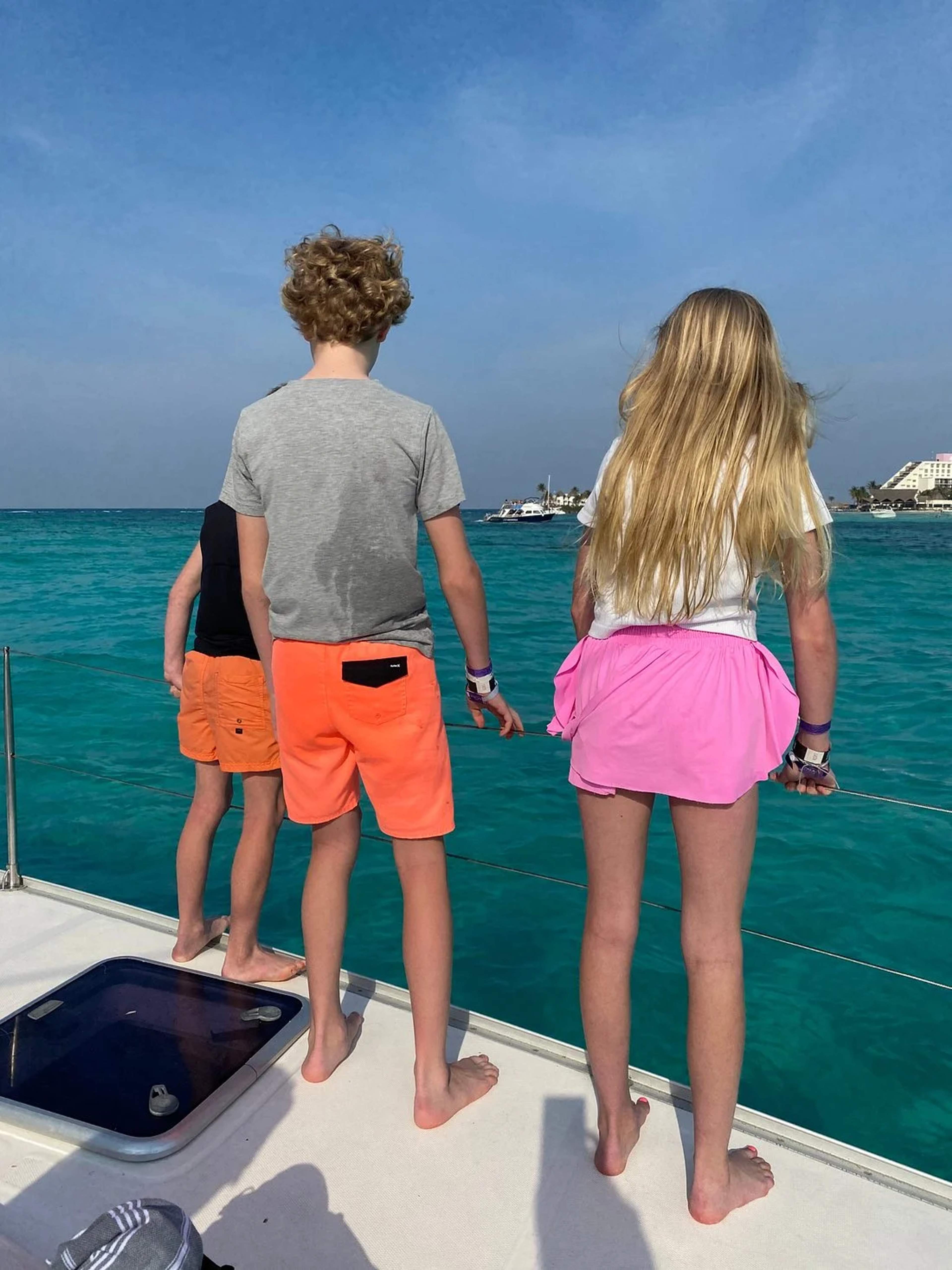 Niños de pie en la cubierta de un bote mirando las aguas turquesas de Cancún en un día soleado.