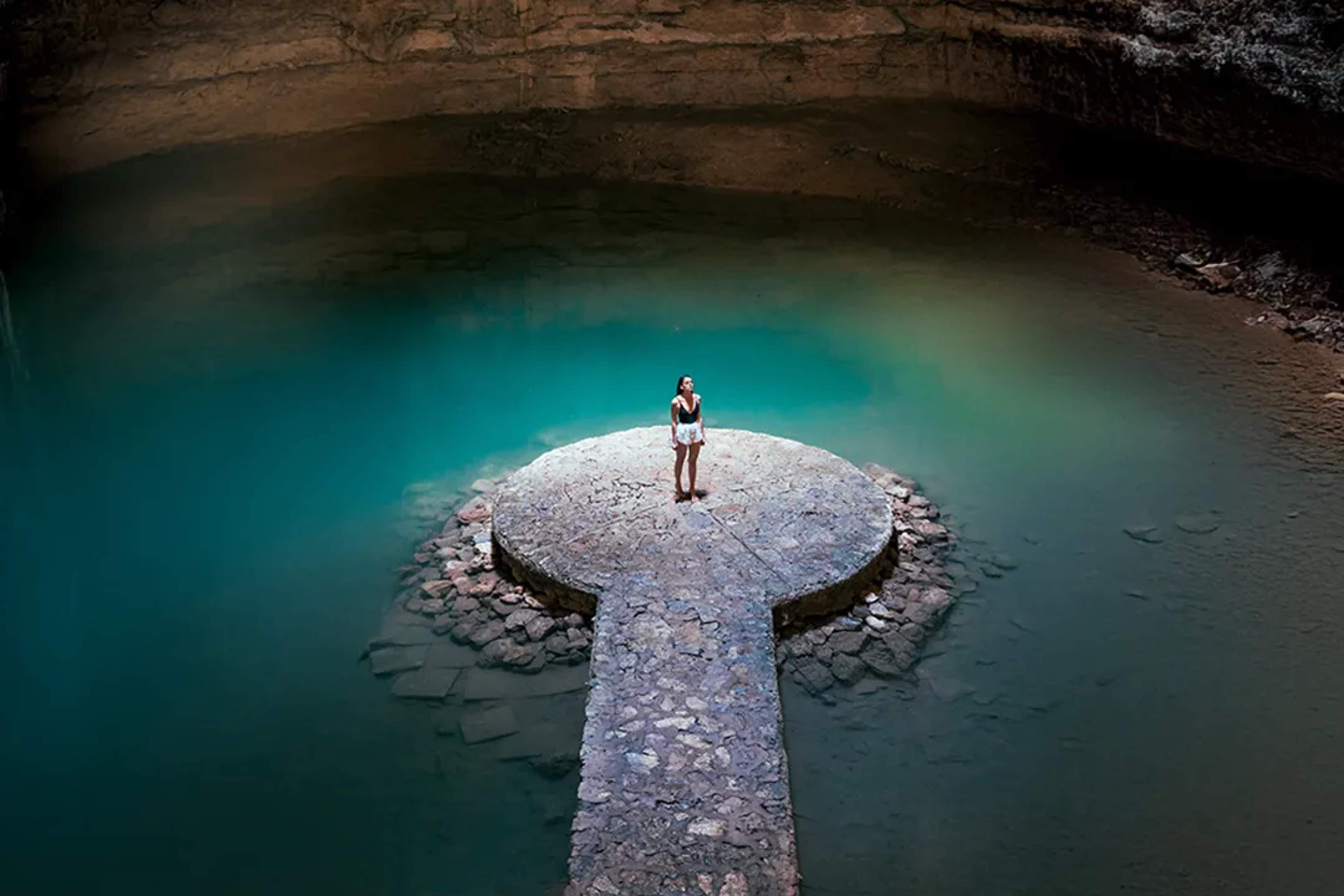 Mujer de pie en plataforma de piedra dentro de un cenote maya de aguas turquesa