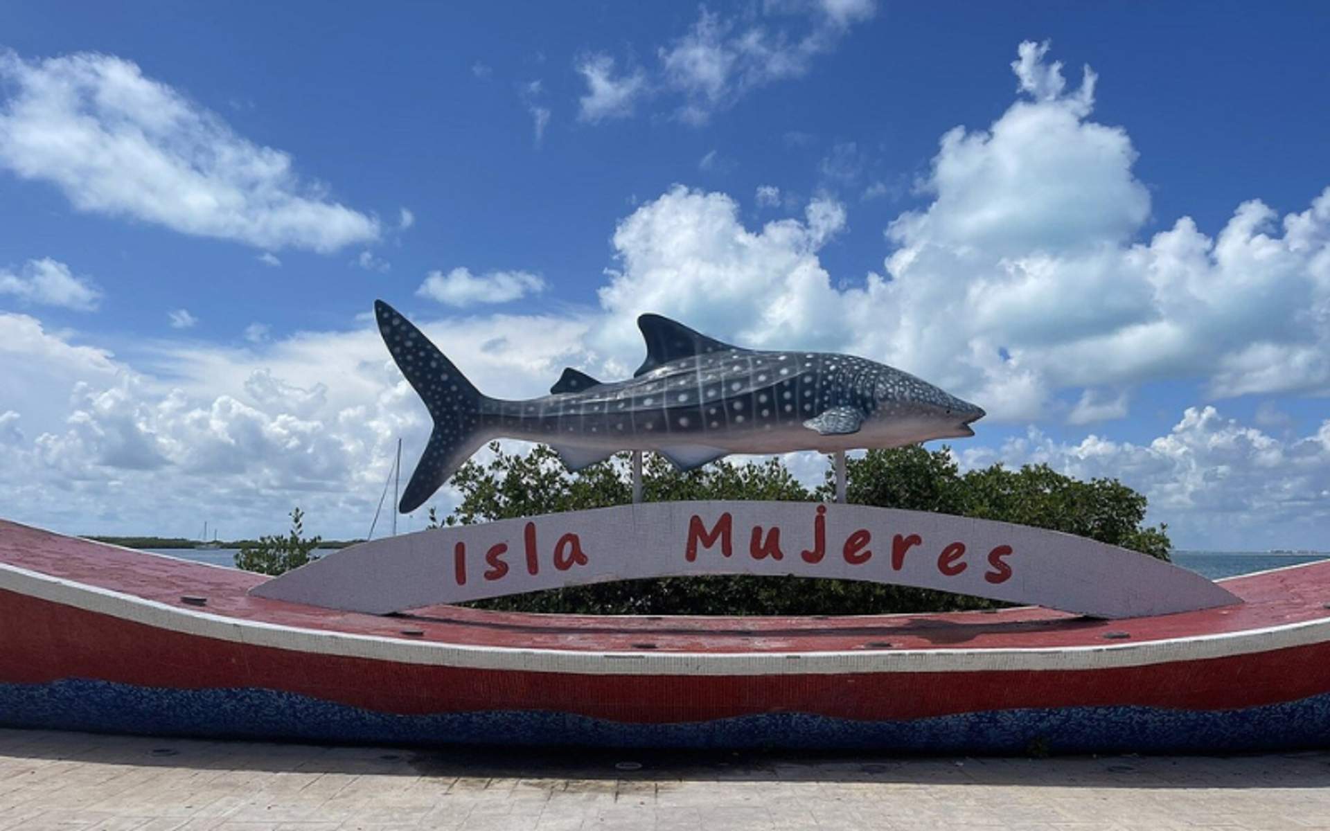 Whale shark statue with "Isla Mujeres" sign under a blue sky with clouds.