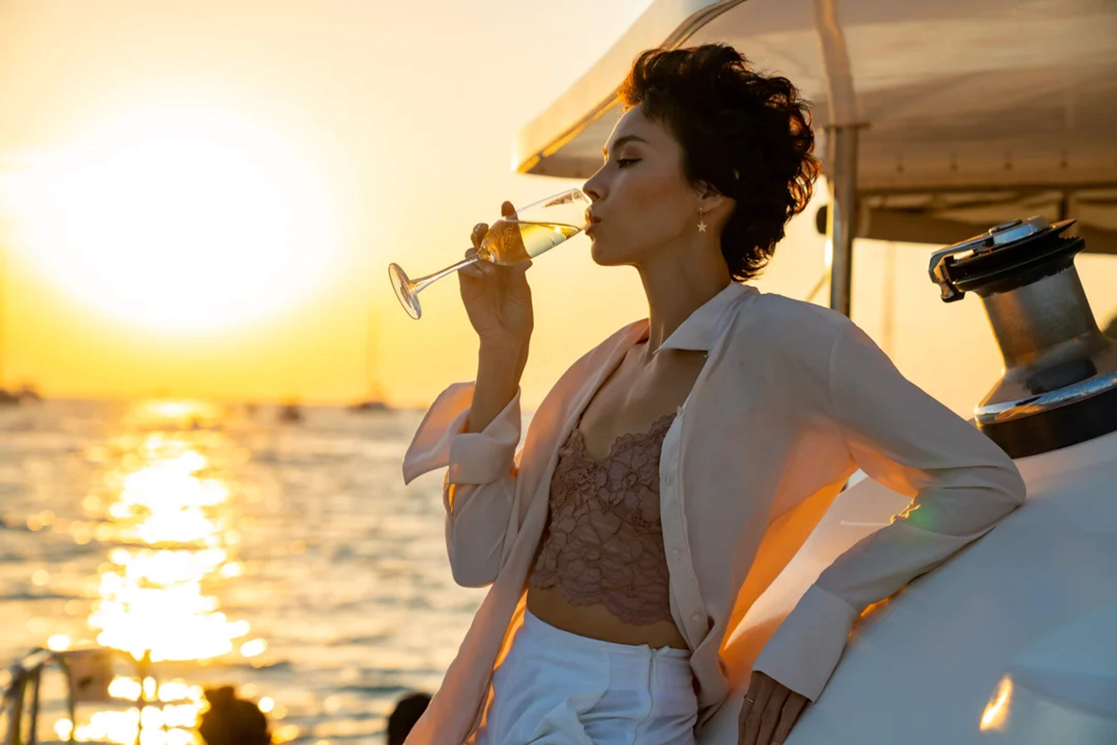 Mujer disfruta una copa de champaña en un yate al atardecer, rodeada de reflejos dorados del mar.