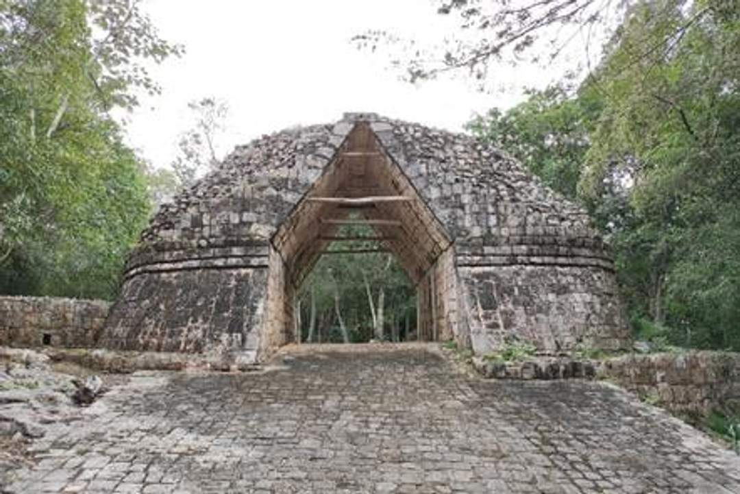 A photograph of the arch in the Initial Series Group at Chichen Itza, showcasing a large, ancient stone archway with a pathway leading through it. The structure is surrounded by lush greenery and partially overcast skies.