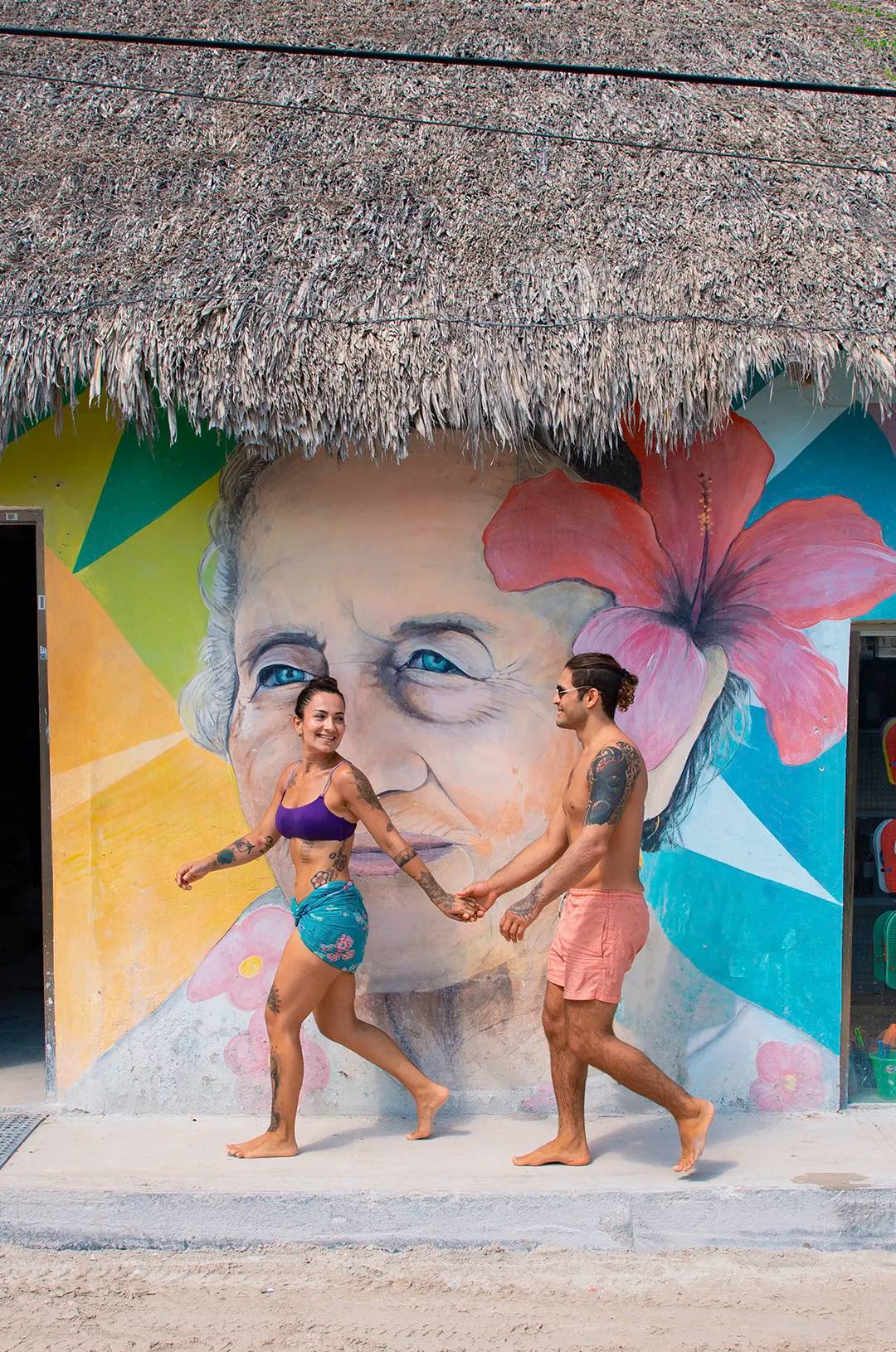 Couple in swimwear holding hands, walking past a mural of an elderly woman's face with a flower in her hair