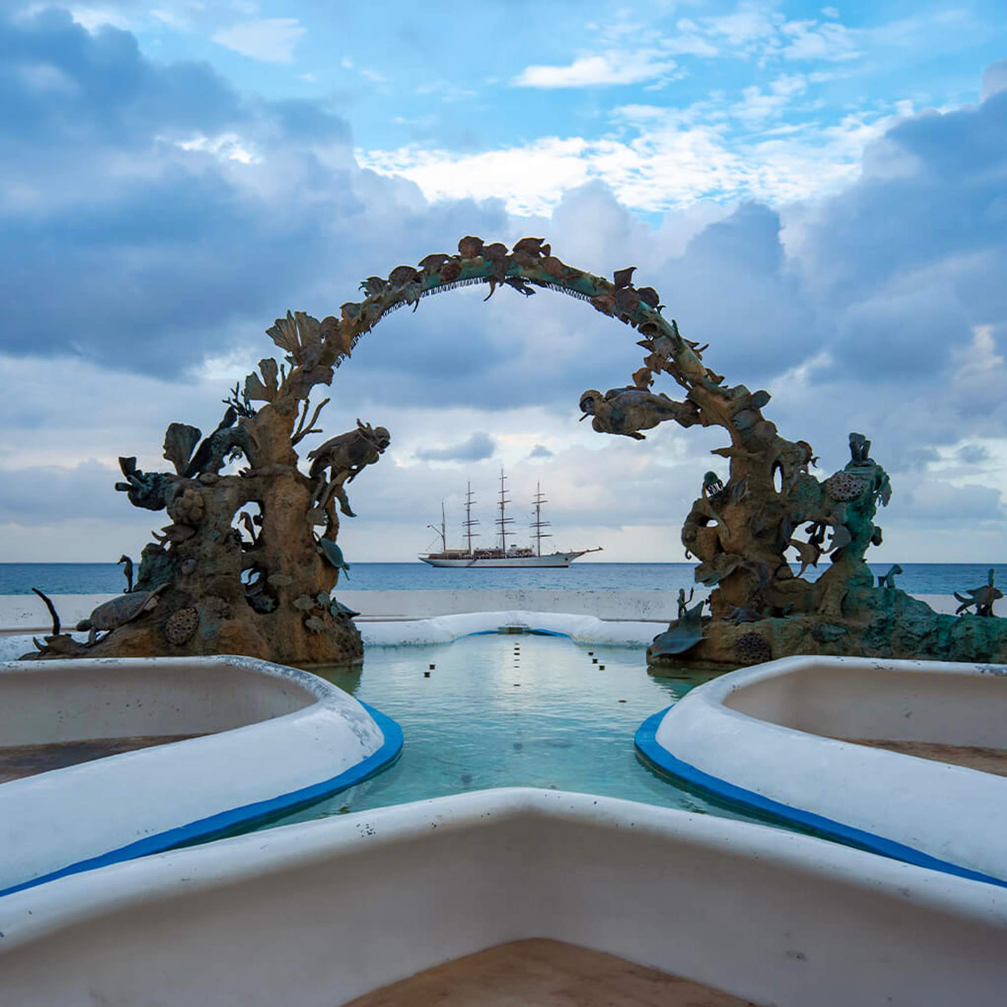 A beautiful ship seen through a unique, intricate arch sculpture by the sea in Cozumel, creating a picturesque scene.