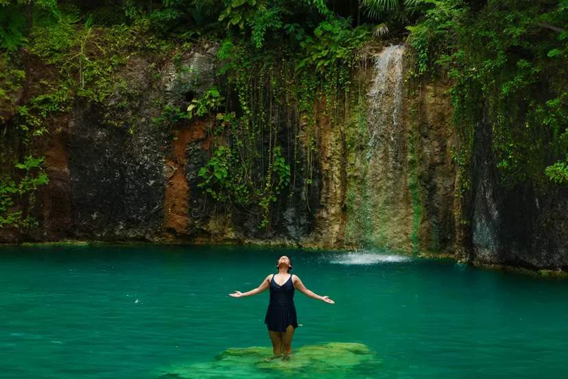 Mujer disfrutando bajo una cascada en una piscina natural de agua turquesa rodeada de exuberante vegetación del Caribe.
