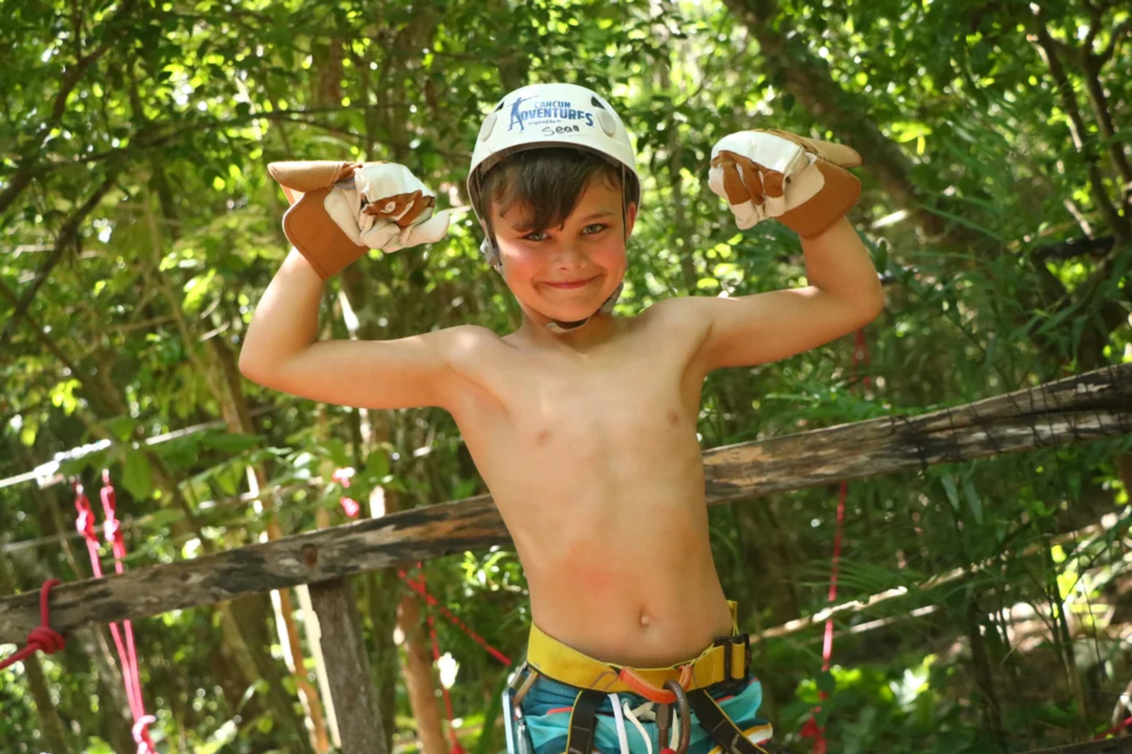 Niño sonriente muestra sus músculos en una aventura en la selva, con casco y equipo de seguridad.