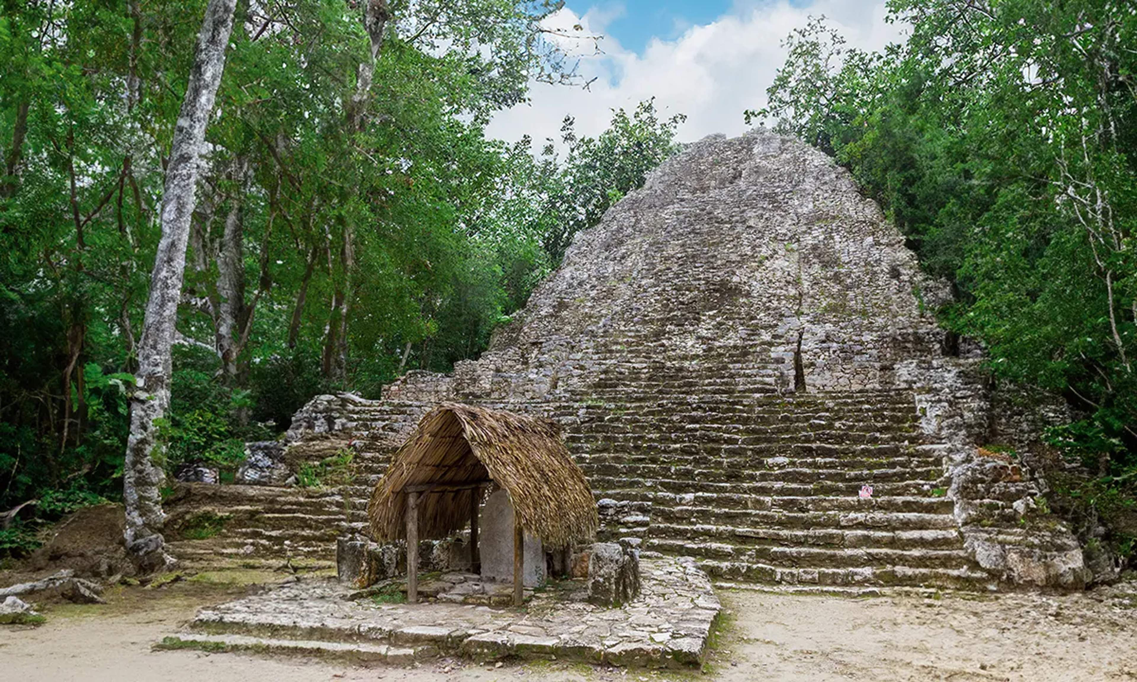 Pirámide antigua de piedra con una pequeña choza de techo de palma en su base, rodeada de selva y un cielo despejado.