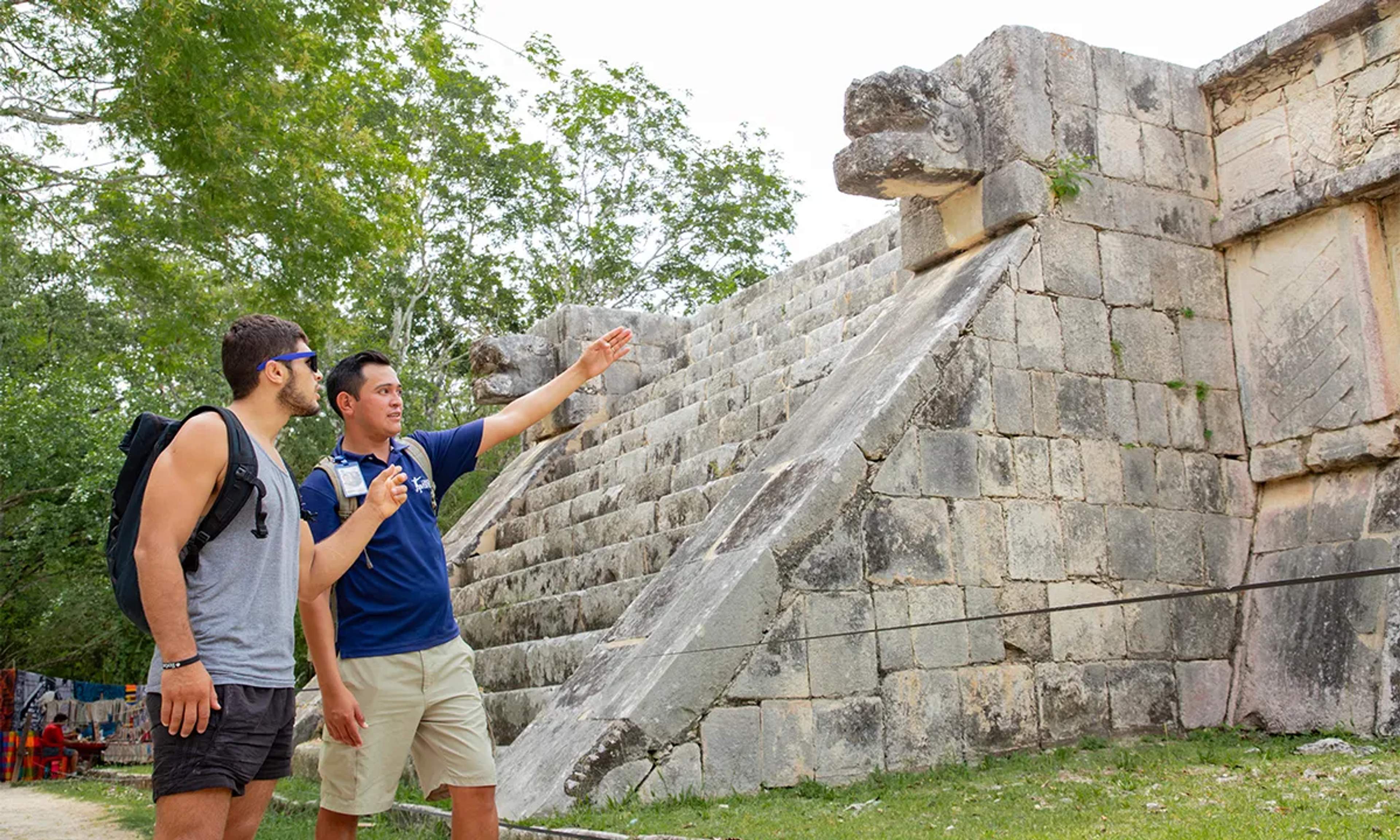 Tres turistas con mochilas están frente al Templo de Kukulcán en Chichén Itzá, México, con plantas verdes en primer plano.