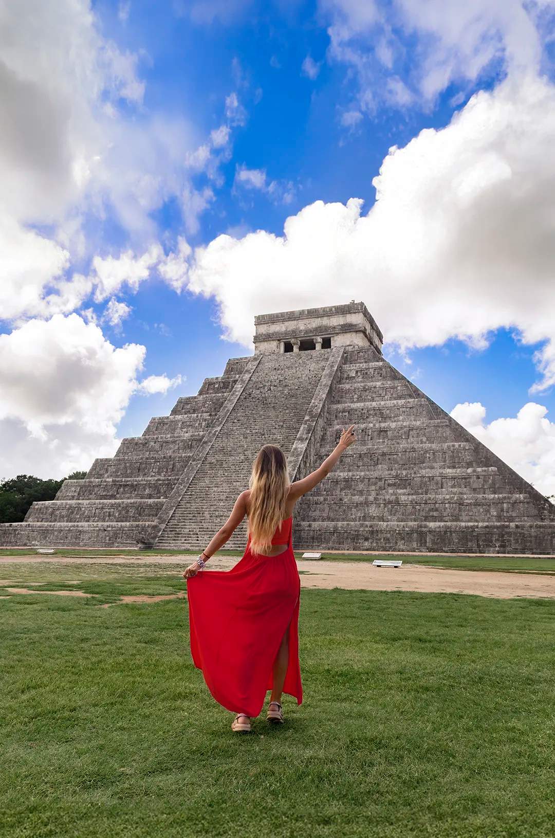 Woman in a red dress, walking towards the Chichen Itza pyramid under a vibrant blue sky with scattered clouds.
