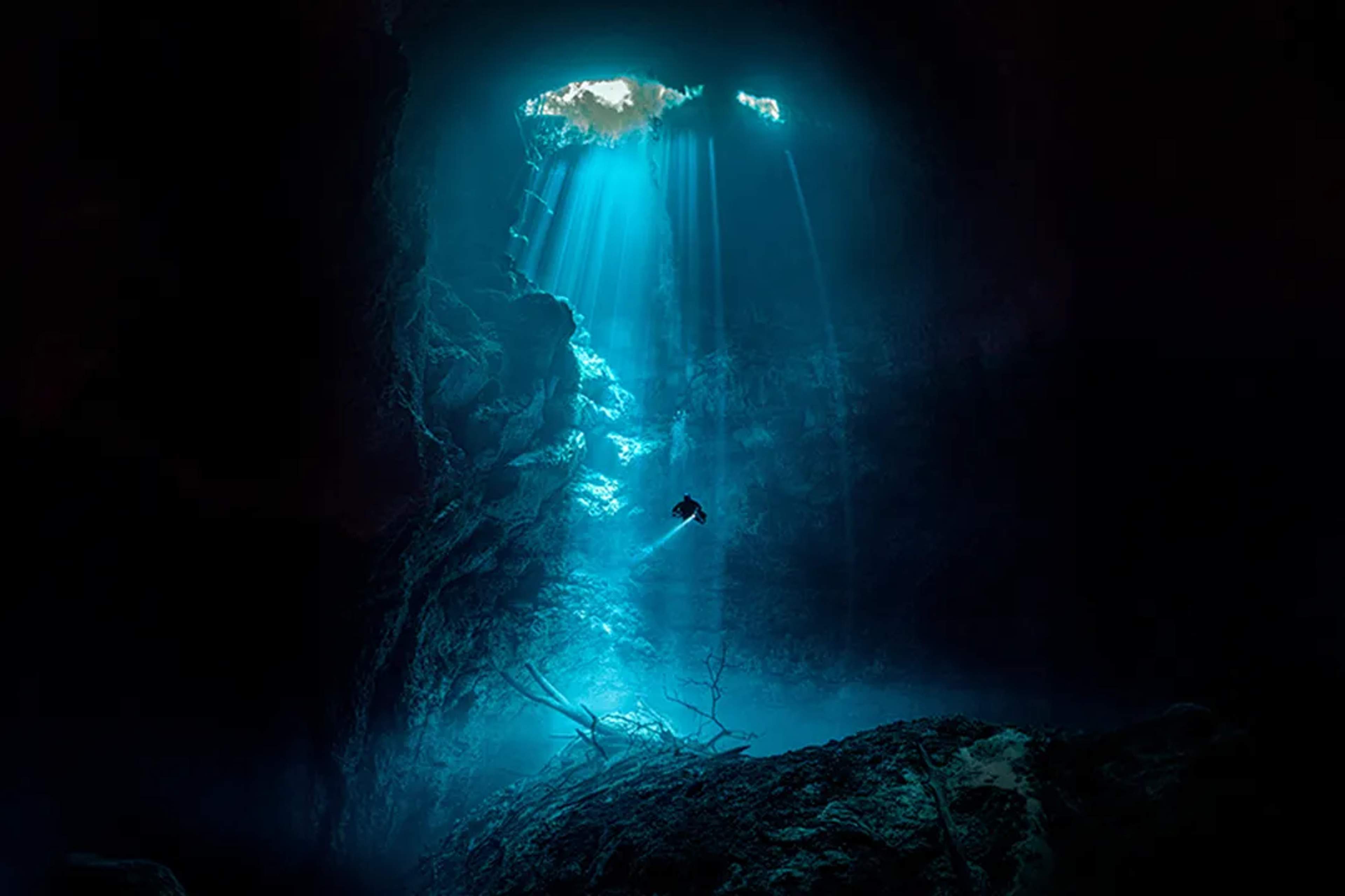 Buzo explora cenote maya con rayos de luz, aguas azul profundo y formaciones rocosas sagradas