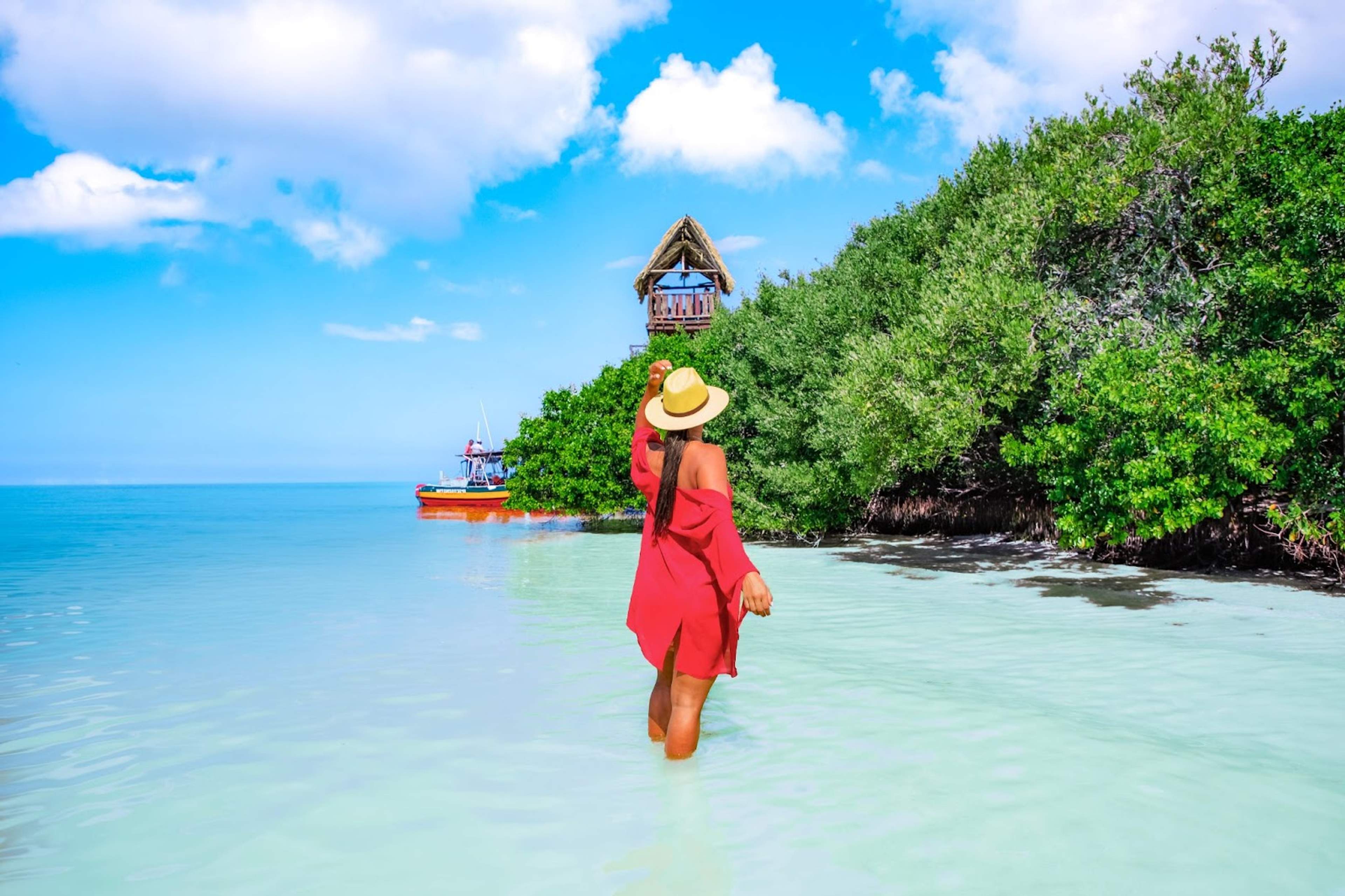 A woman in a red dress and hat wades through clear blue water toward a thatched-roof lookout, with a red boat anchored nearby and lush greenery in the background.