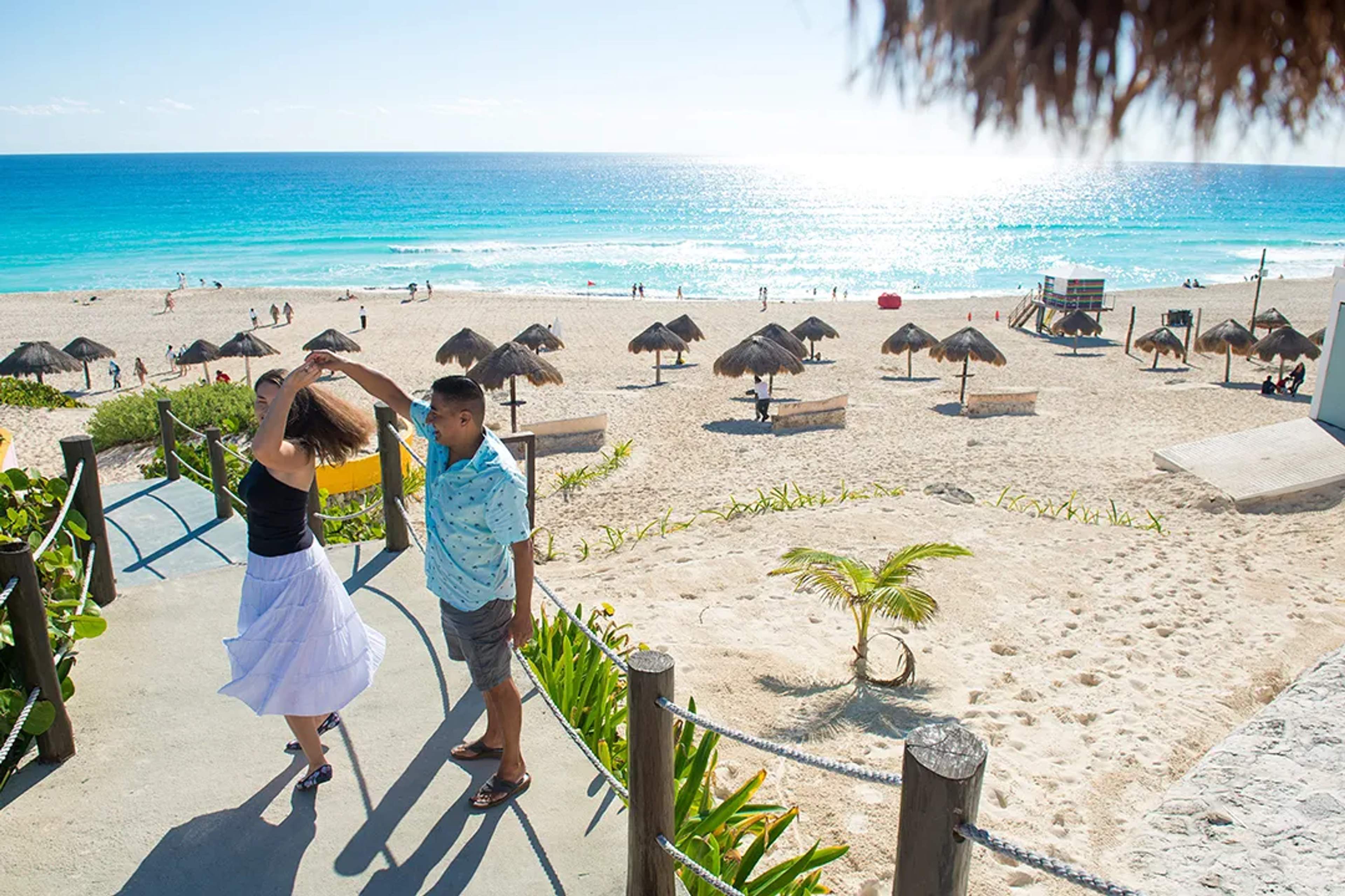 Couple dancing by the beach in Cancun with turquoise ocean, white sand, and palm huts on a sunny day