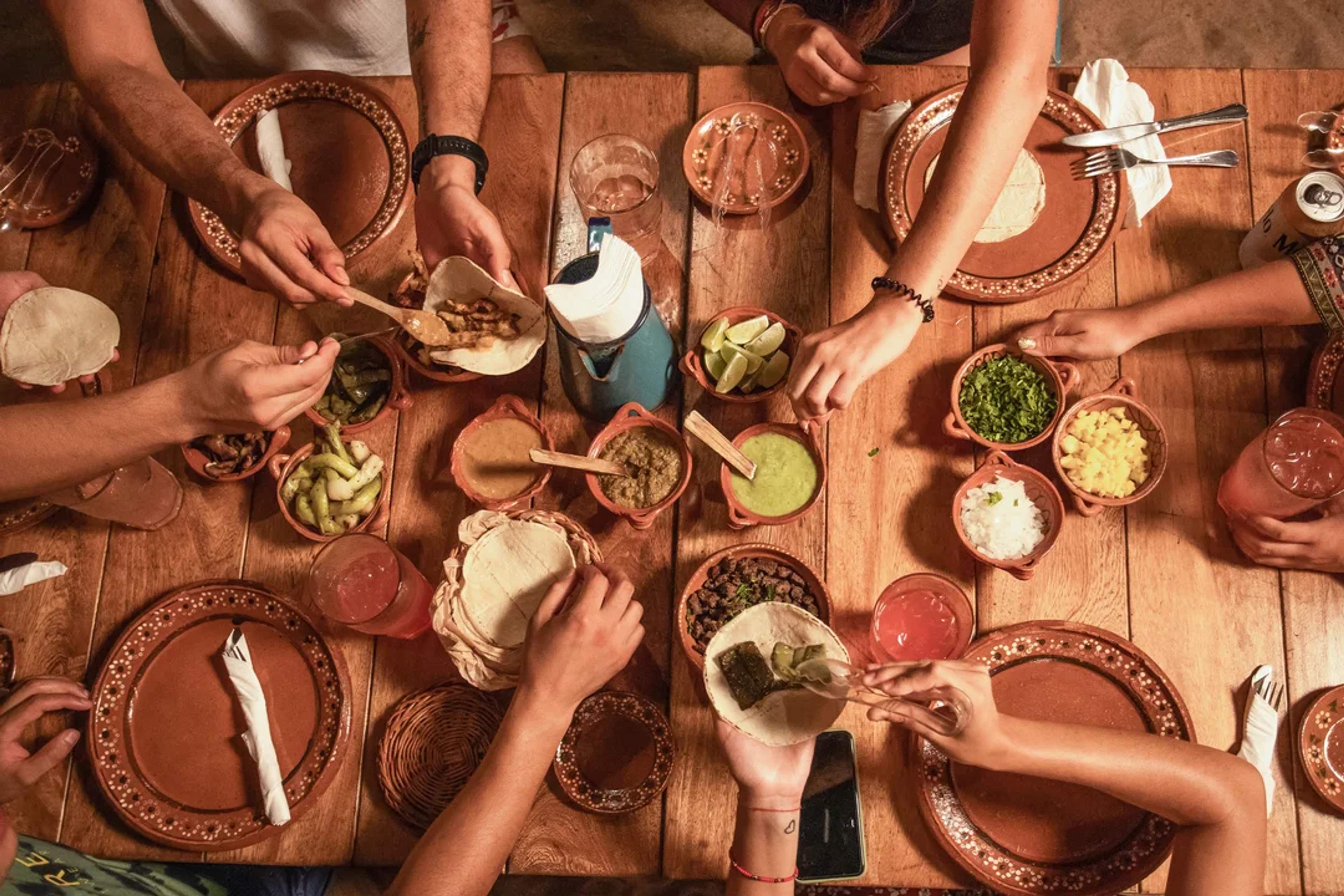 Personas disfrutan preparando tacos alrededor de una mesa llena de tortillas, guisos, salsas y acompañamientos frescos.