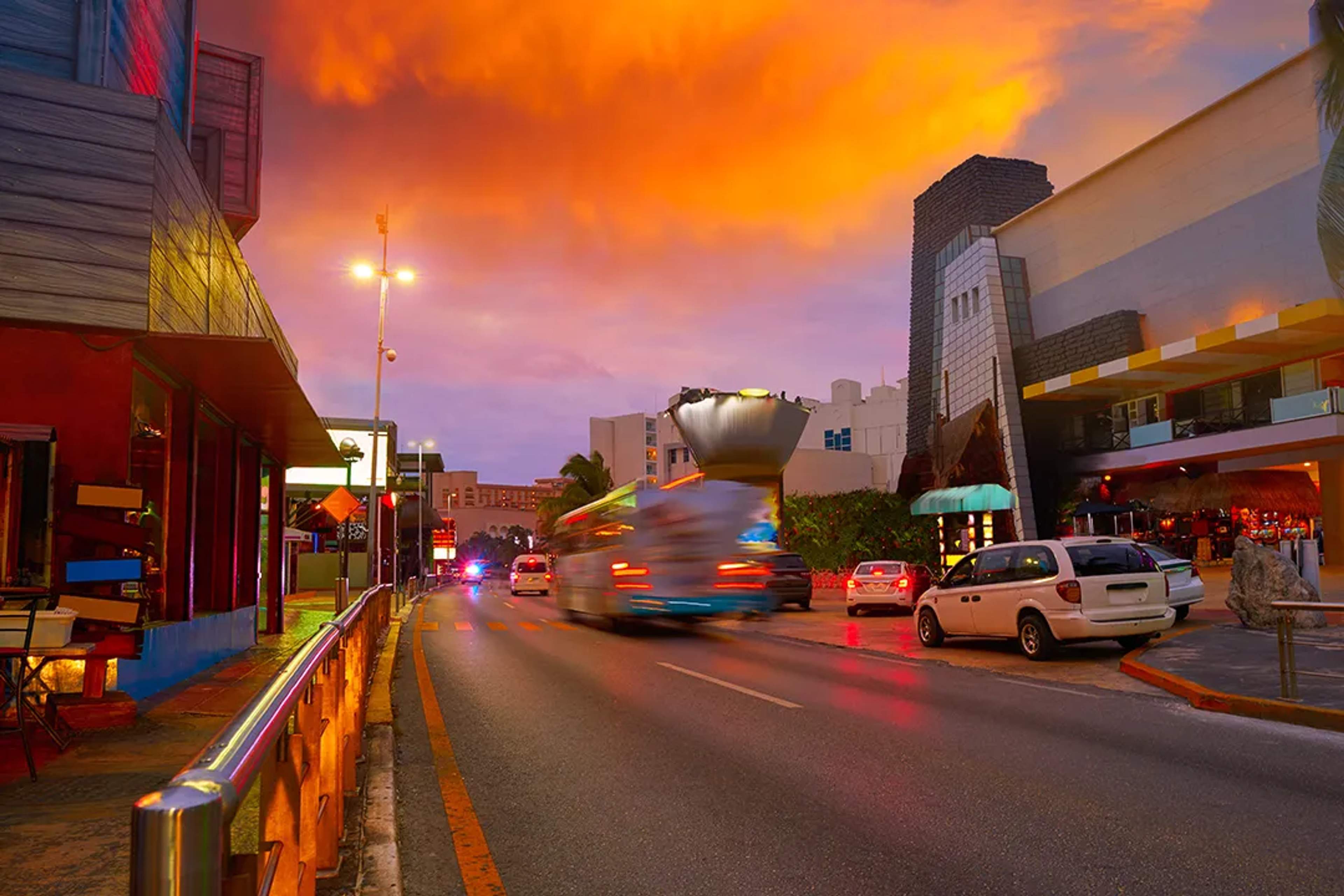 Boulevard de Cancún al atardecer con luces vibrantes, tráfico en movimiento y locales de vida nocturna