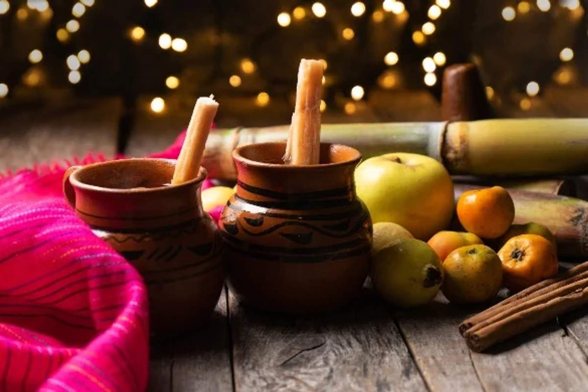 Dos tazas de barro tradicionales mexicanas con bebida caliente, rodeadas de caña de azúcar, manzanas, tejocotes, ramas de canela y un paño rosa vibrante, con luces festivas de fondo.