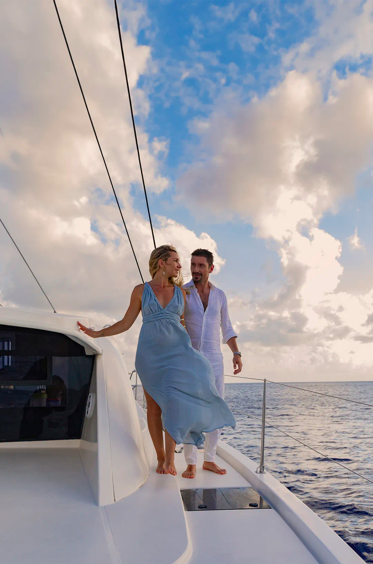 Una pareja camina descalza en un yate al atardecer, con la mujer en un vestido azul y el hombre en ropa blanca.