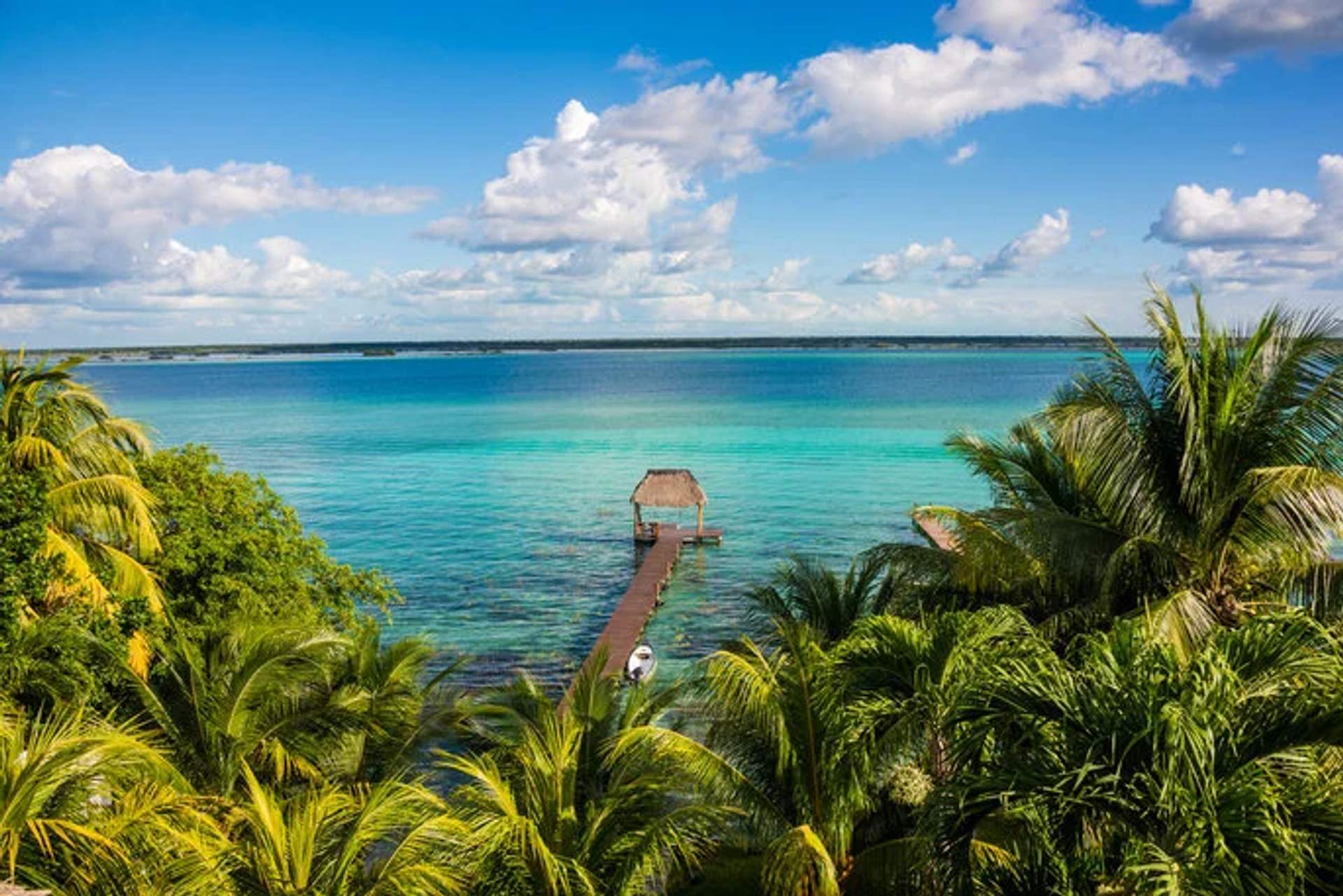 Scenic view of Lake Bacalar with a wooden pier, thatched roof hut, and lush palm trees under a blue sky.